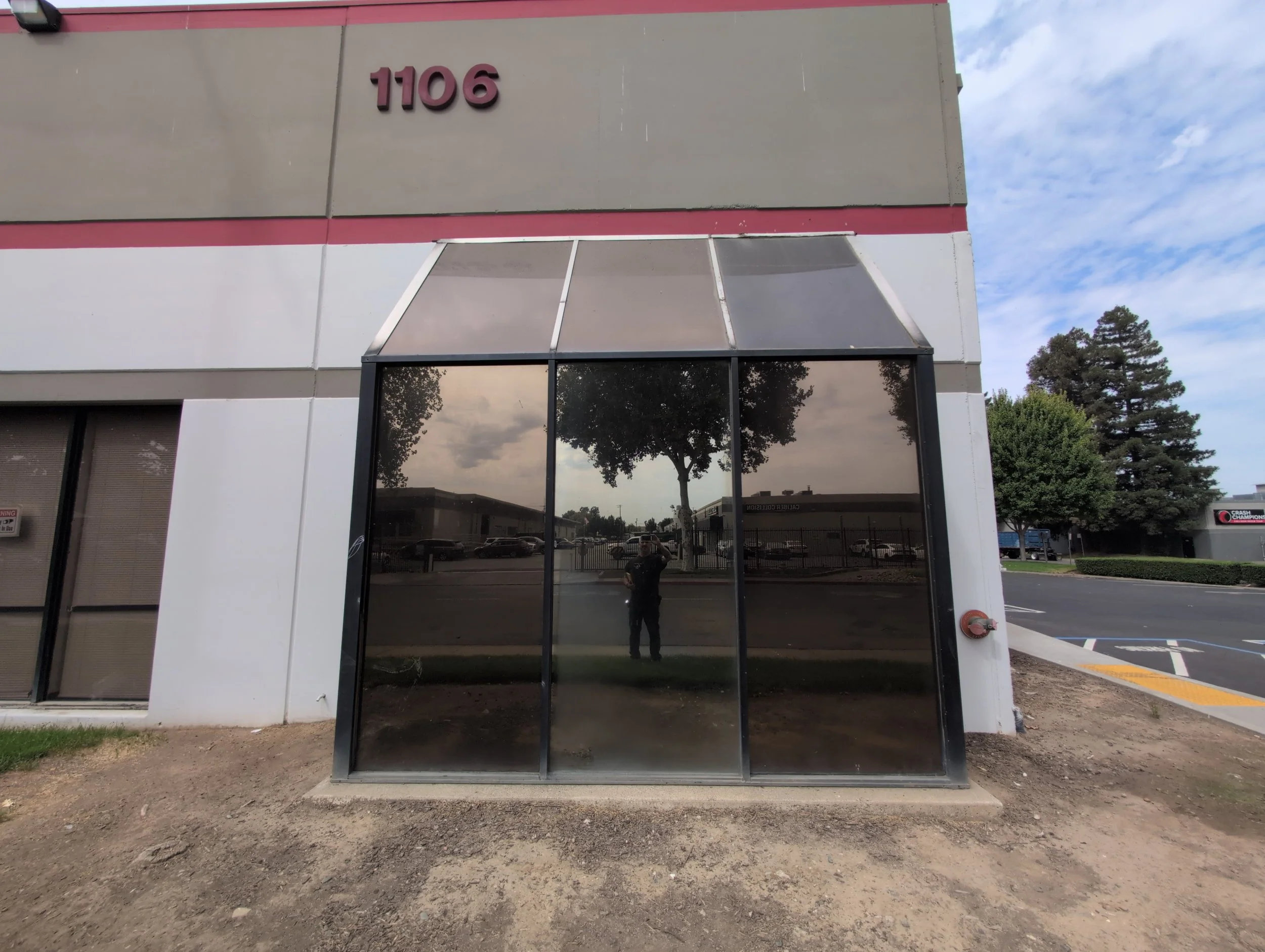 Building exterior with the number 1106 displayed at the top, reflective glass door in the center reflecting trees and the sky, and an ATM visible on the left side. Gravel and dirt ground in the foreground.