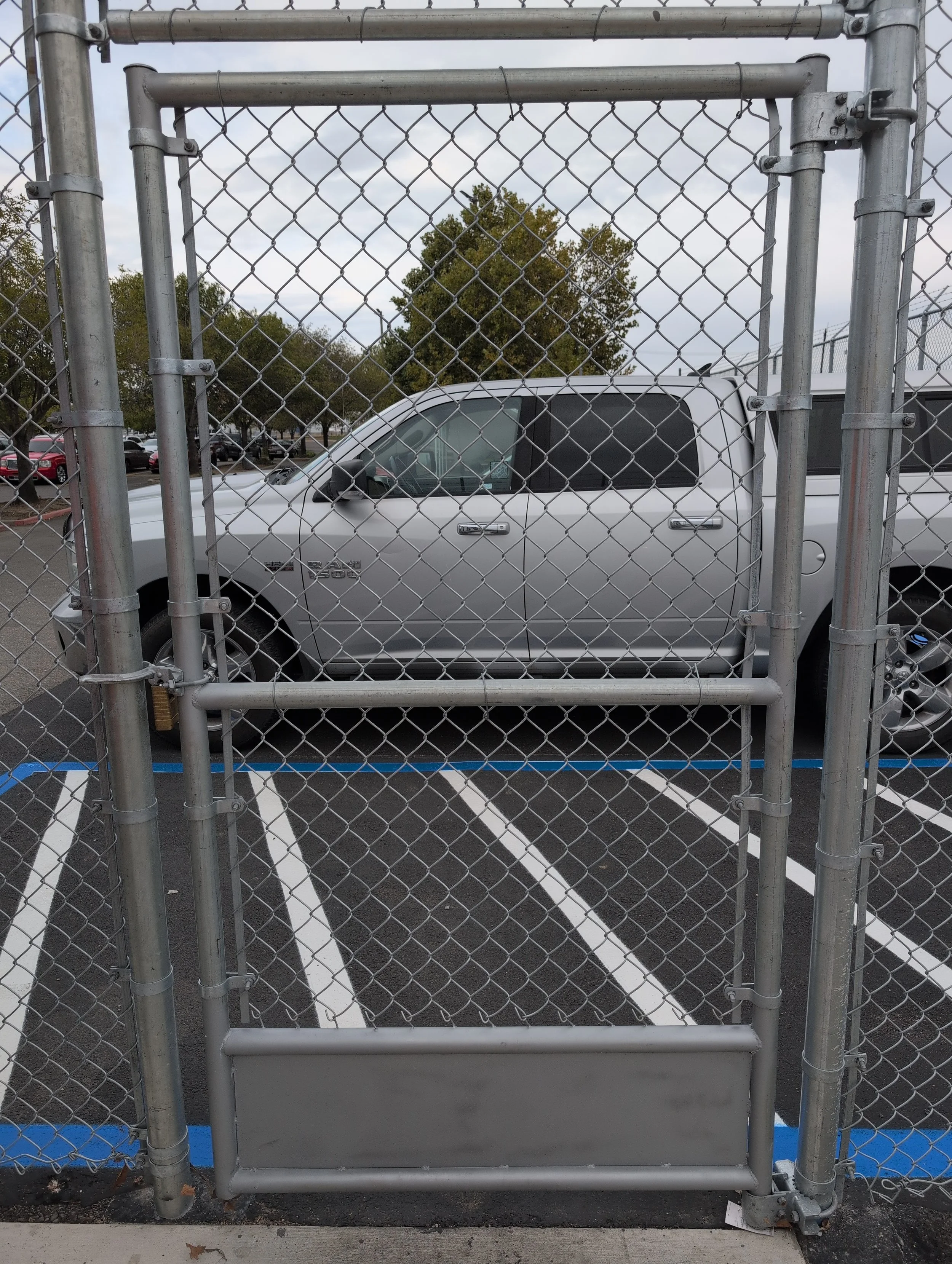 The image shows a silver pickup truck parked behind a chain-link fence with an additional security gate. The parking lot has white parking lines and a few trees in the background with a cloudy sky.