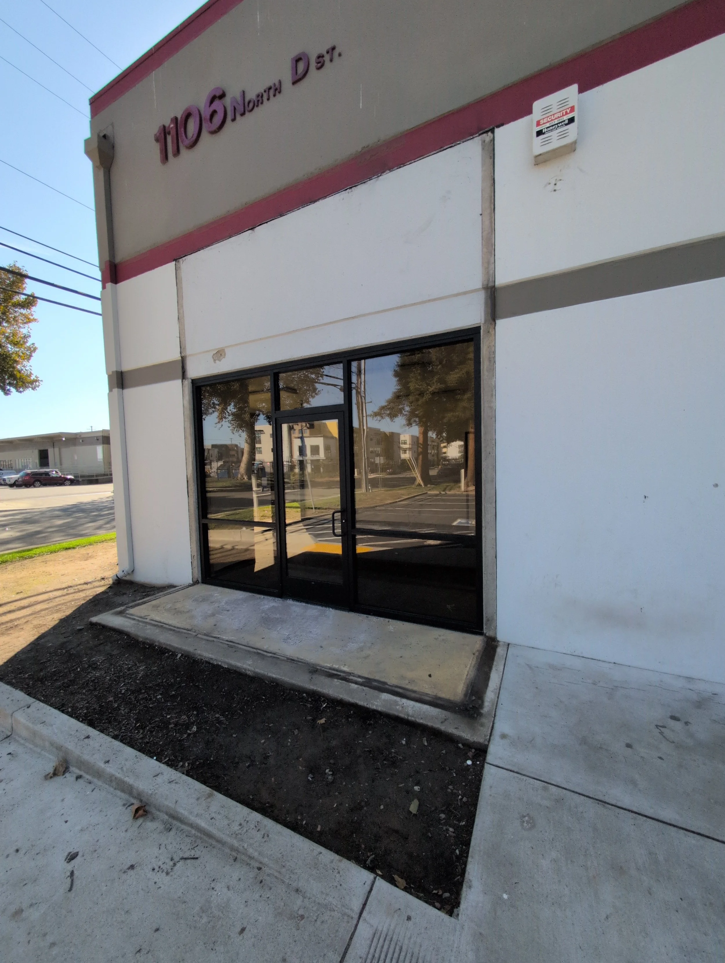 A commercial building with a glass door entrance, unfinished soil in front, and a sign showing the address '1105 North D St.' on the upper corner.