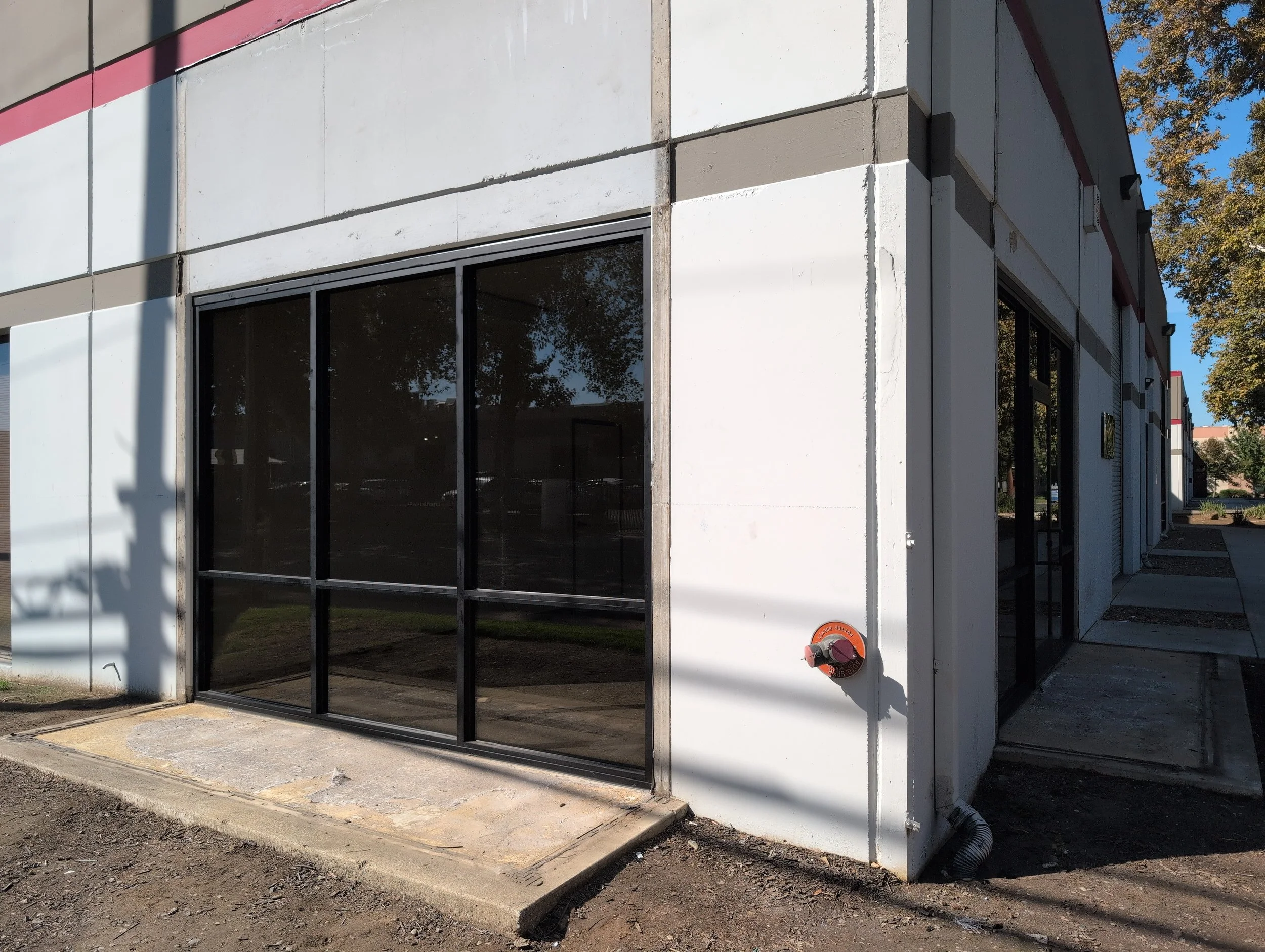 Exterior of a commercial building under construction with large glass windows and white panels, construction materials on the ground, and a black drainage pipe on the right side.