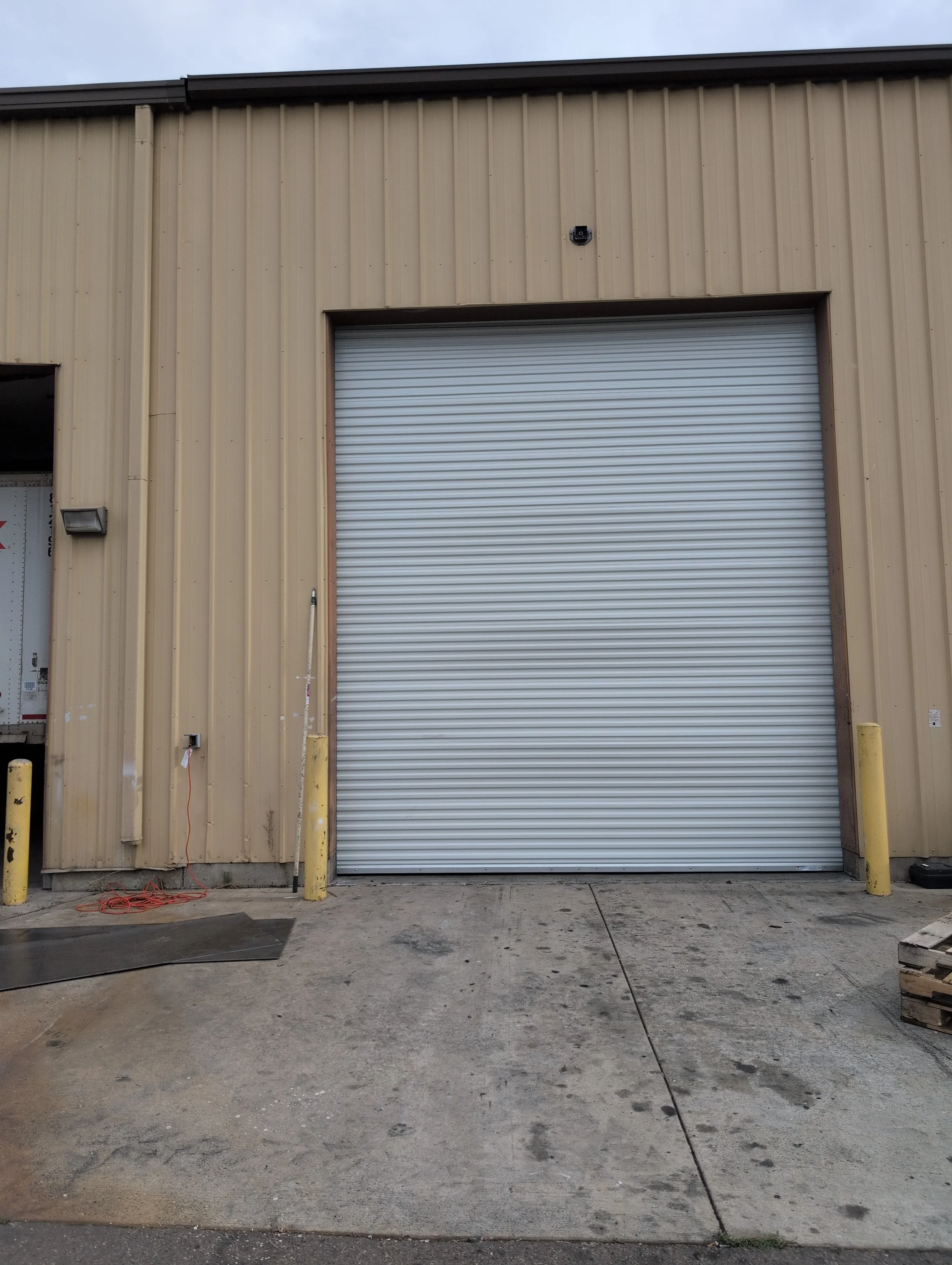 The exterior of a beige industrial building with a large closed metal roll-up door. There is a security camera above the door, and two yellow bollards in front of it. The ground is concrete with some stains and debris.