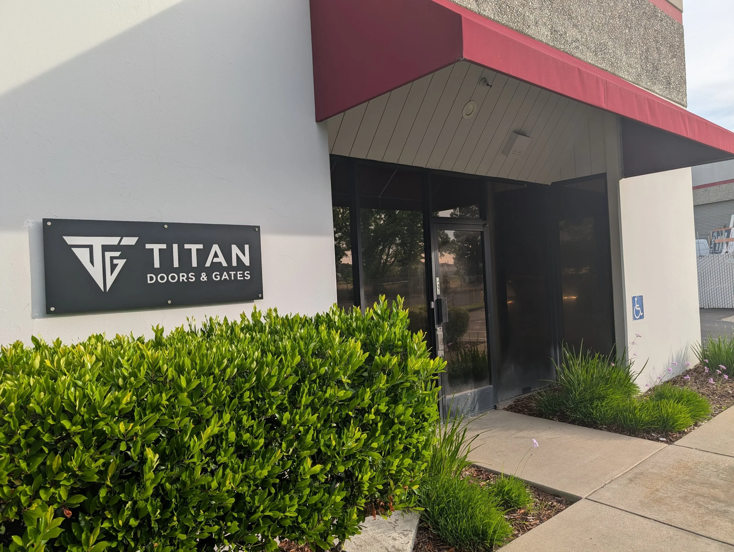 Building entrance with sign that says "Titan Doors & Gates" and greenery in front.