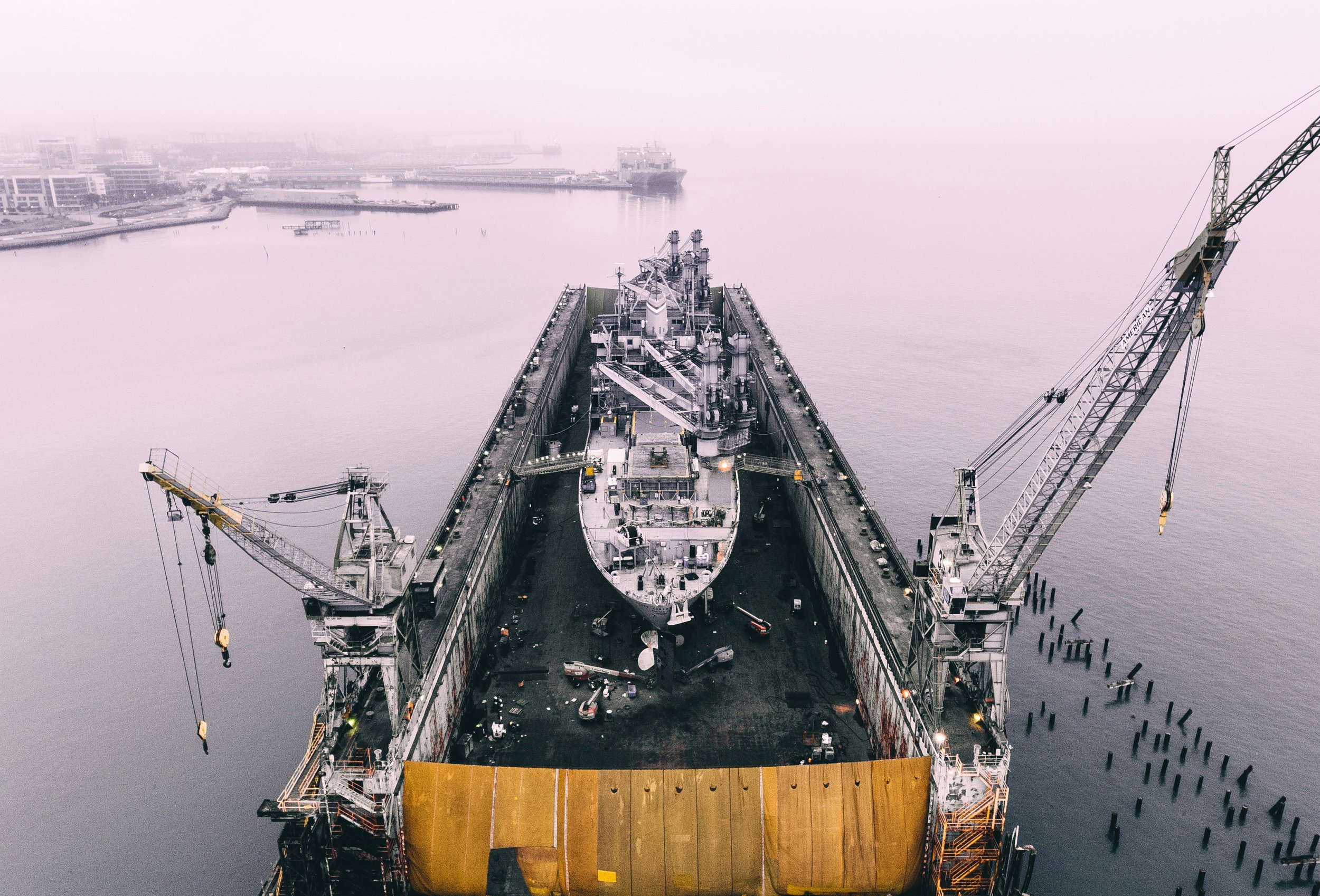 Aerial view of a large ship inside a dry dock at a harbor, with cranes and industrial buildings in background.