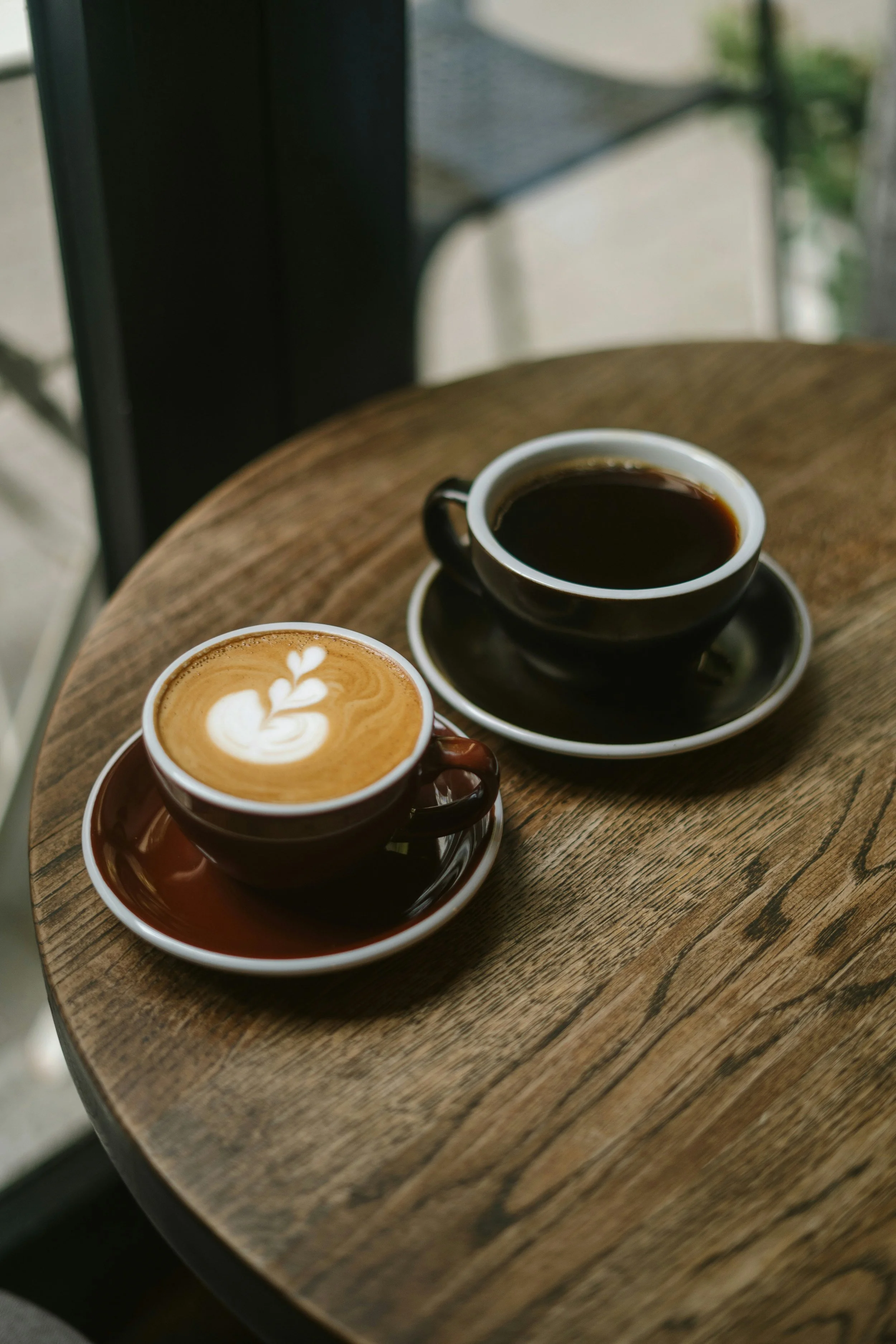 Two cups of coffee, one with latte art and the other black, on a wooden table near a window.