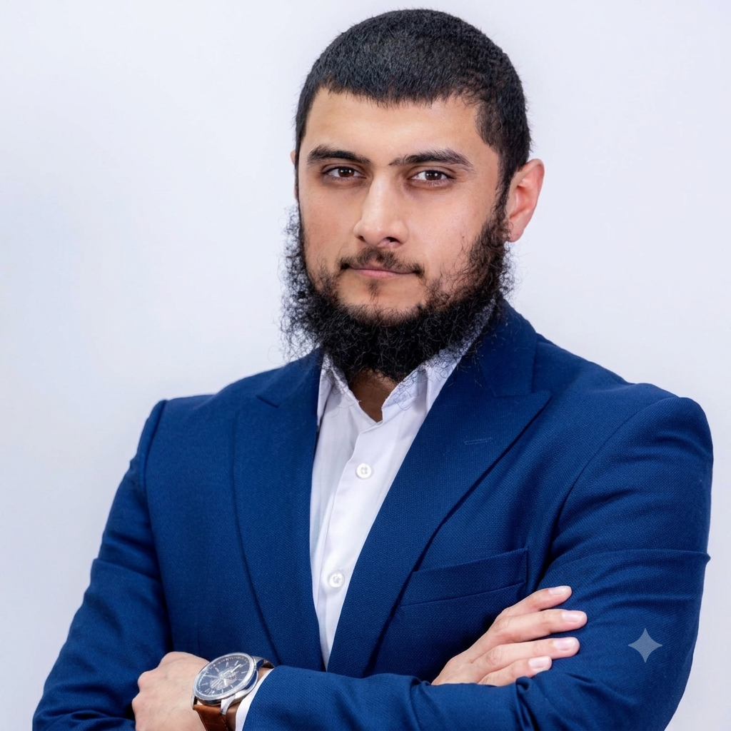 A young man with a beard and short hair wearing a blue suit and white shirt, standing with arms crossed against a plain light background.