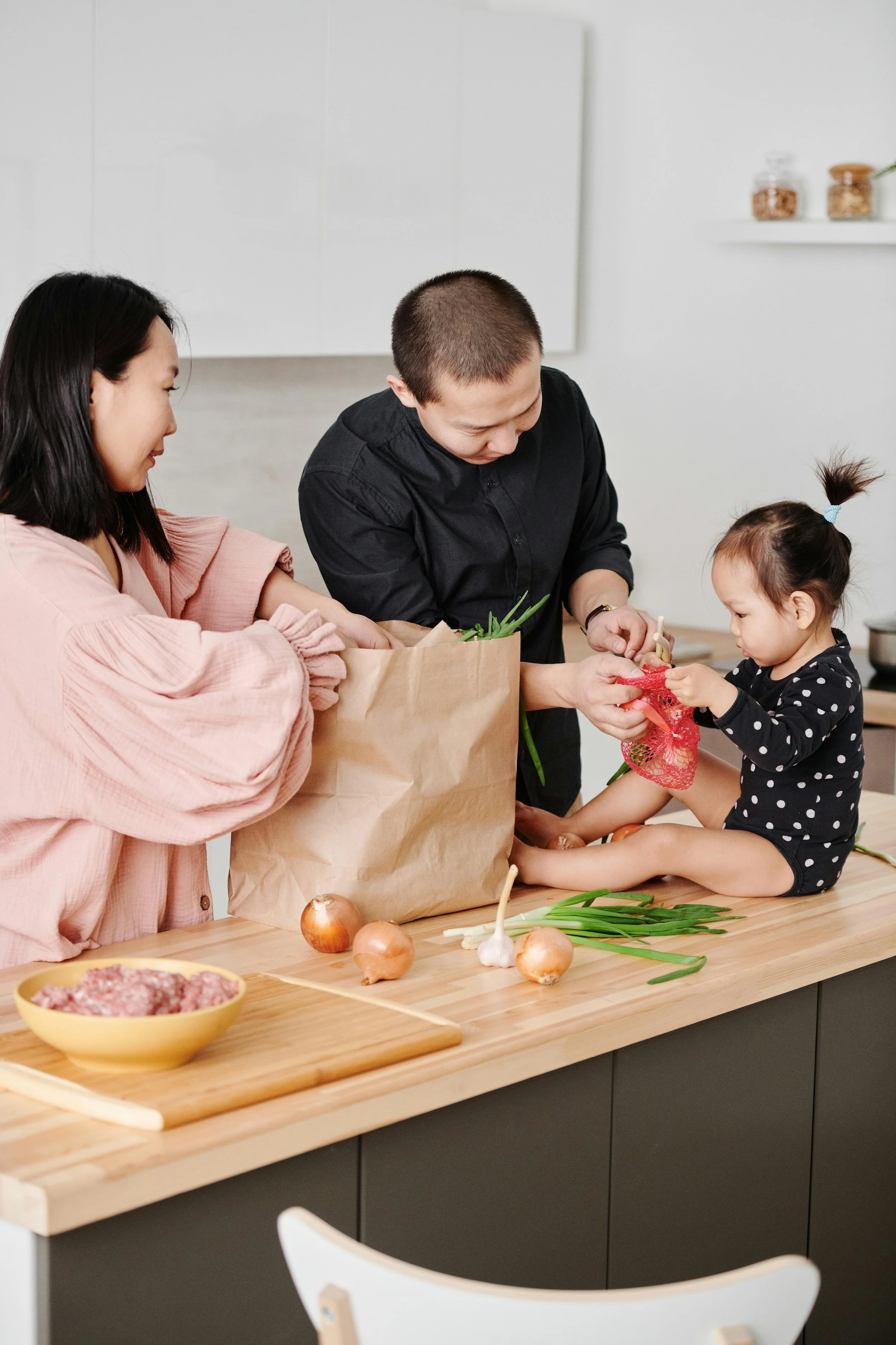 Three people in a kitchen preparing a meal together, with onions, green onions, and ground meat on the counter.