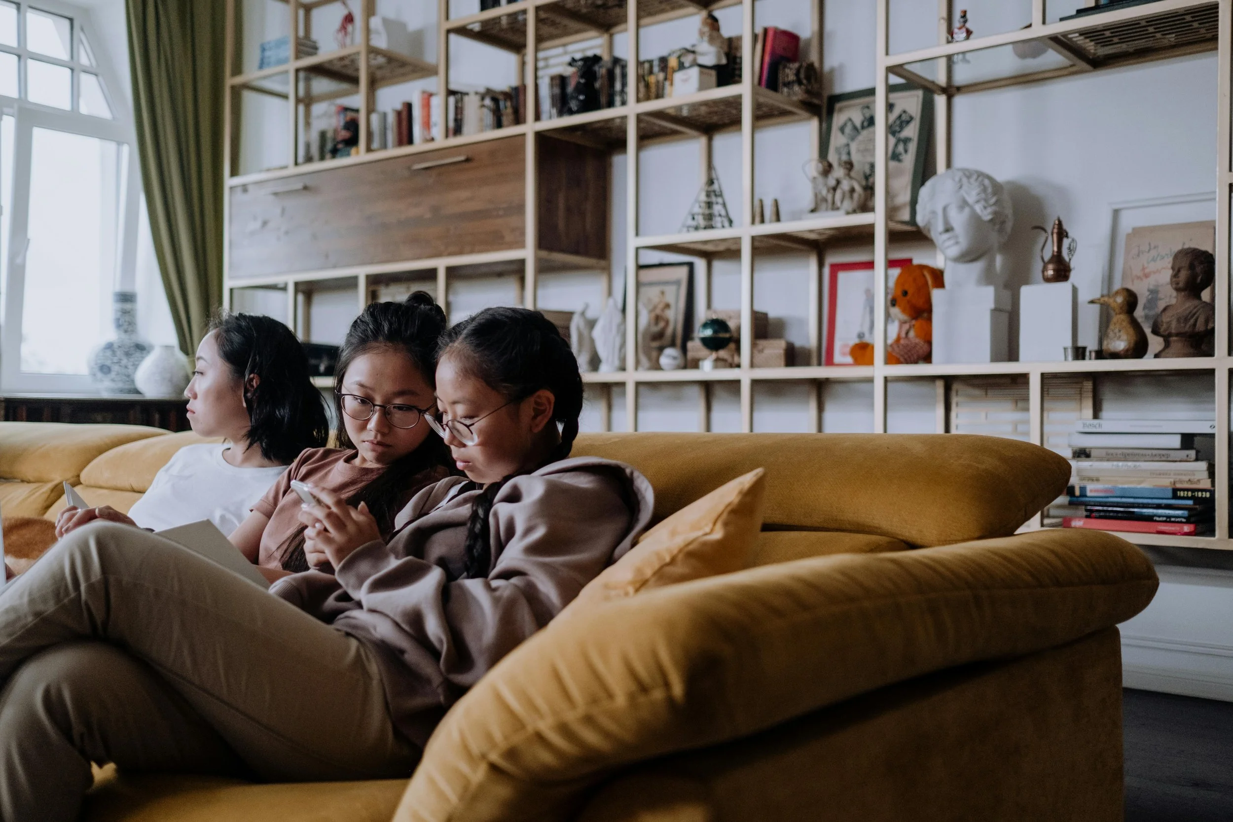 Three young women sitting on a yellow sofa, looking at their phones, with a large wooden shelving unit filled with books and decorative objects behind them in a well-lit room with a window and green curtains.