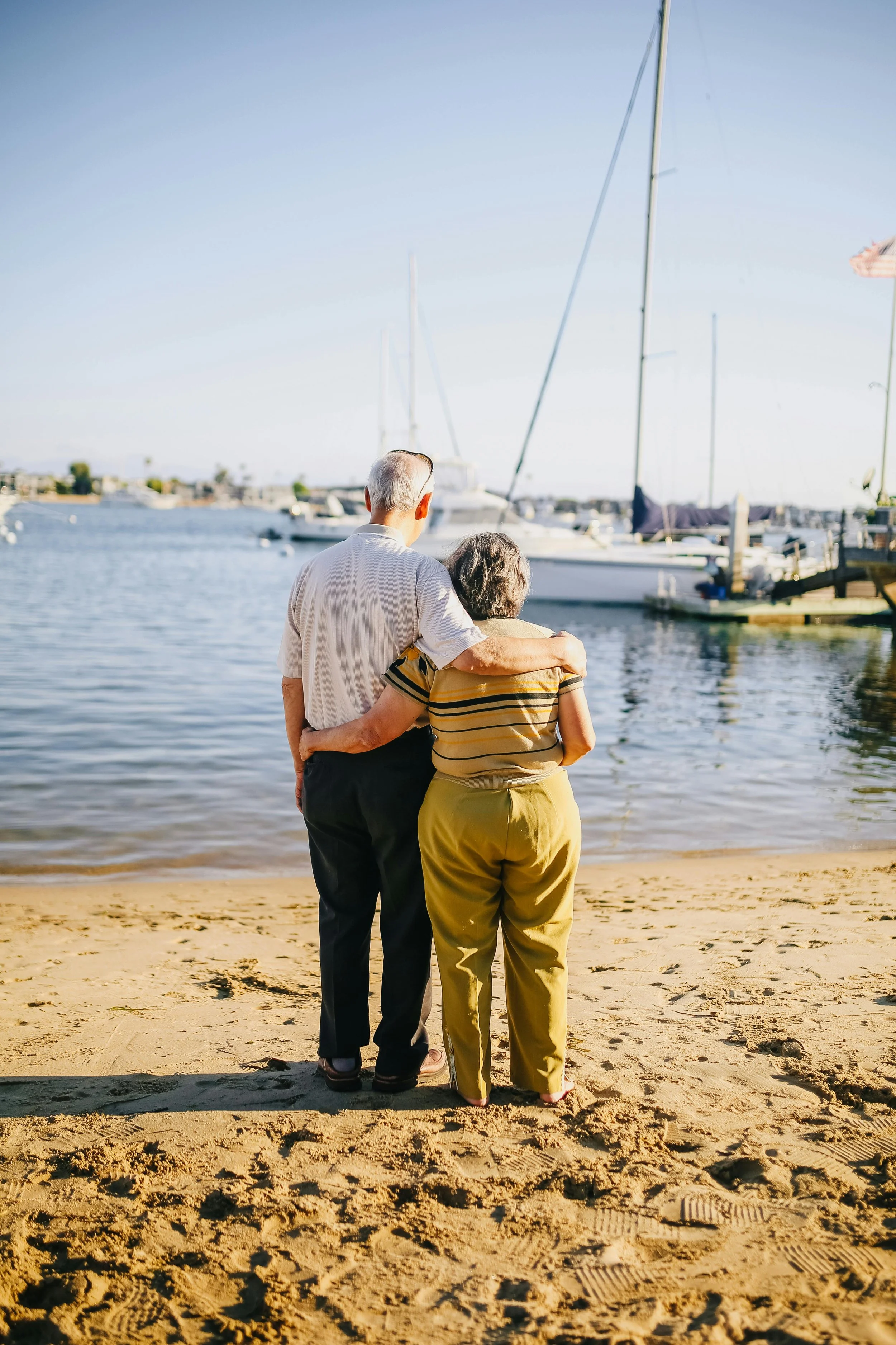 A senior man and woman standing on a sandy beach, gazing at boats docked in a harbor, with the man having his arms around the woman's shoulders.
