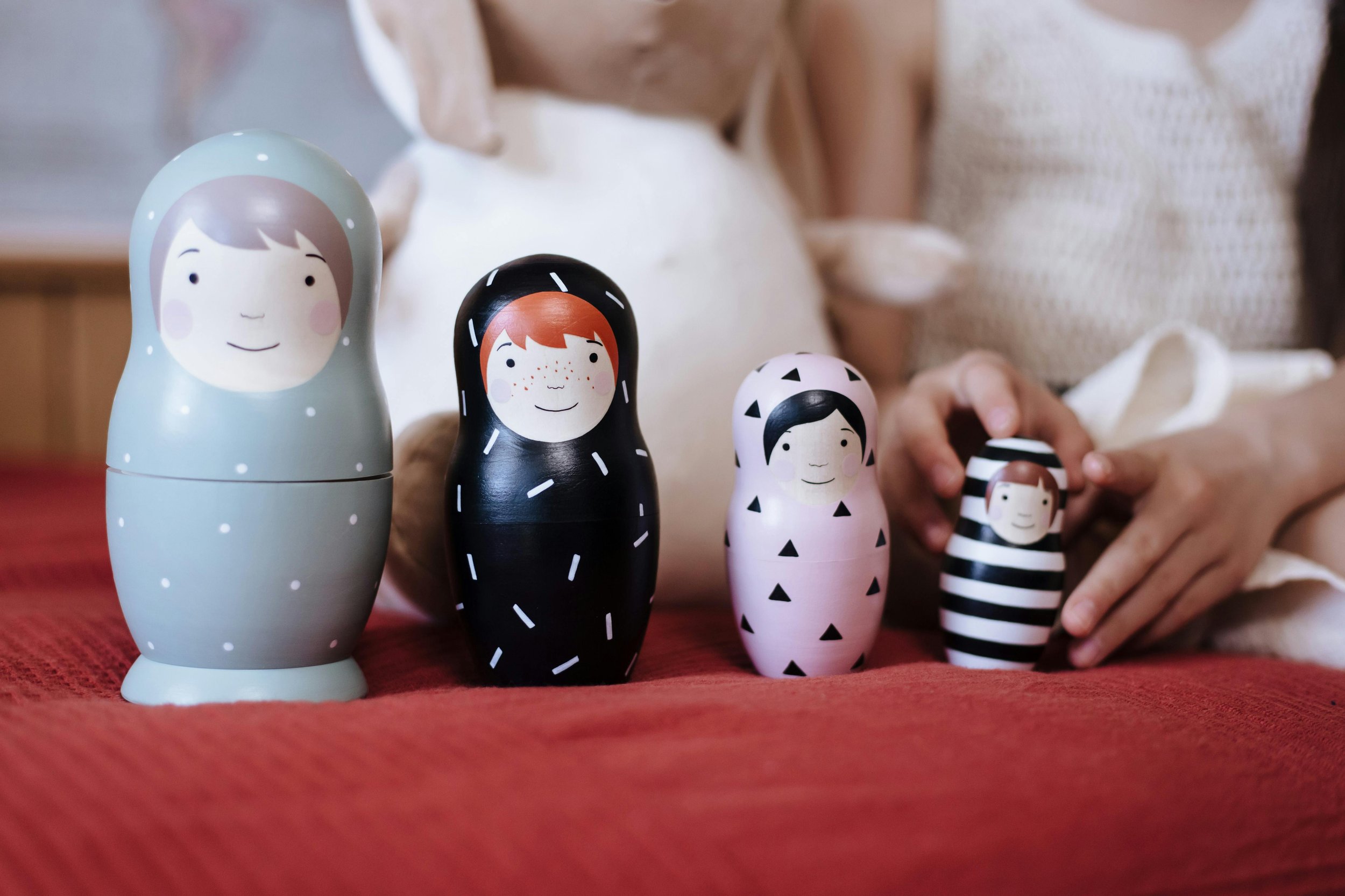 A collection of five painted nesting dolls with different faces and designs on a red surface, with a person in the background reaching toward them.