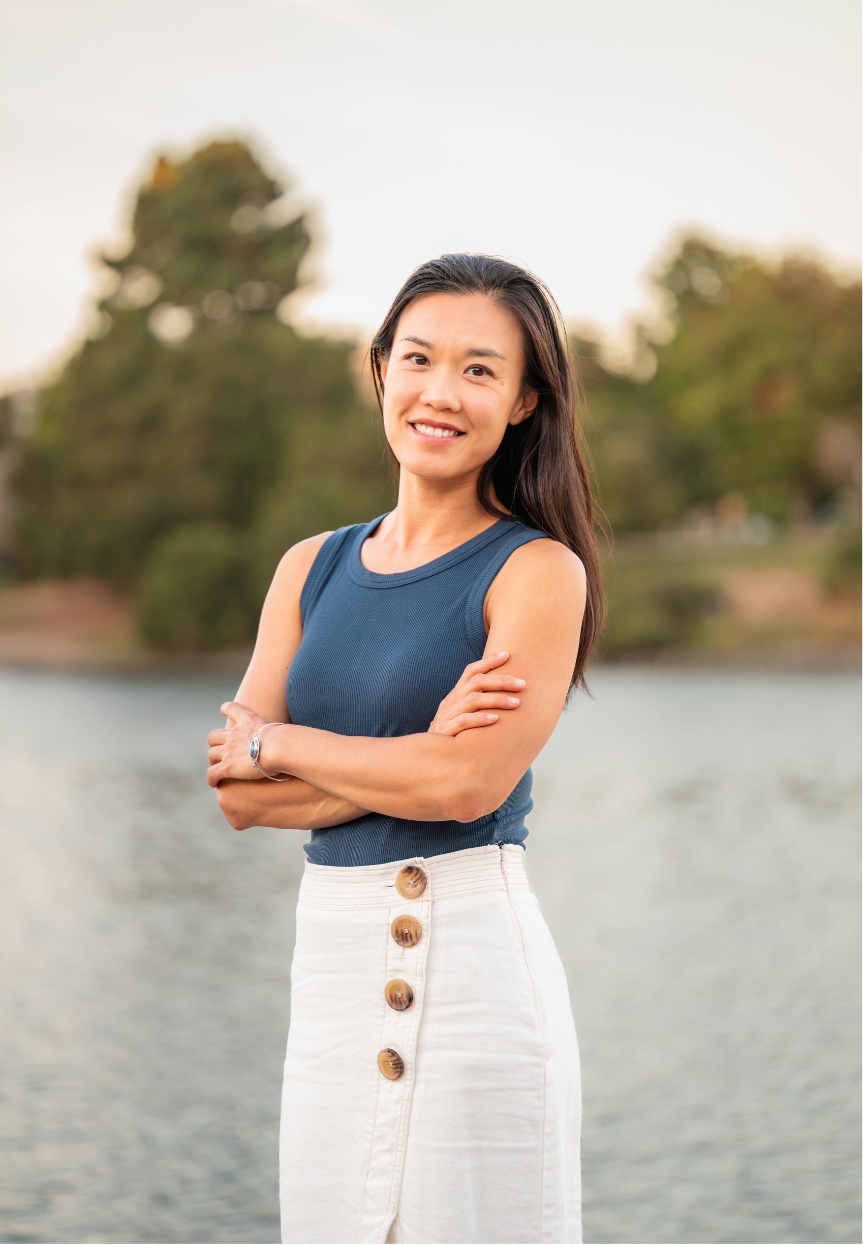 Laura Bai, an Asian American therapist in Oakland, CA, standing outdoors near a body of water, smiling with arms crossed, wearing a blue sleeveless top and a white skirt with large buttons.