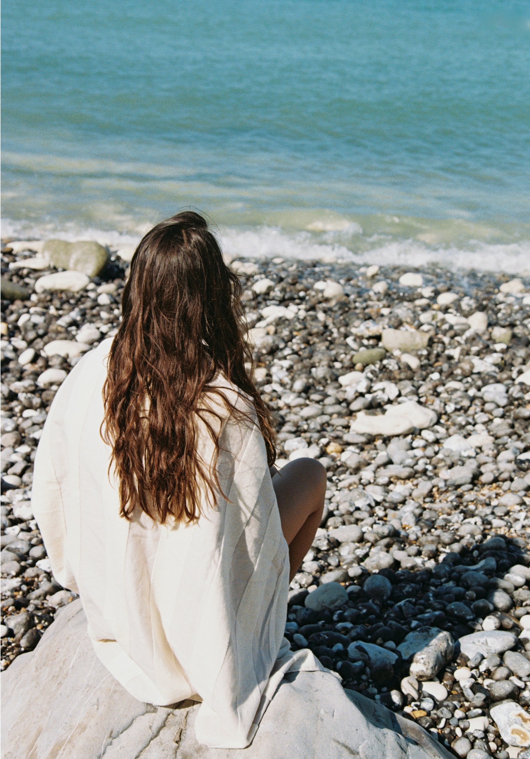 A woman with long brown hair sitting on a rock at a pebble beach looking at the ocean.