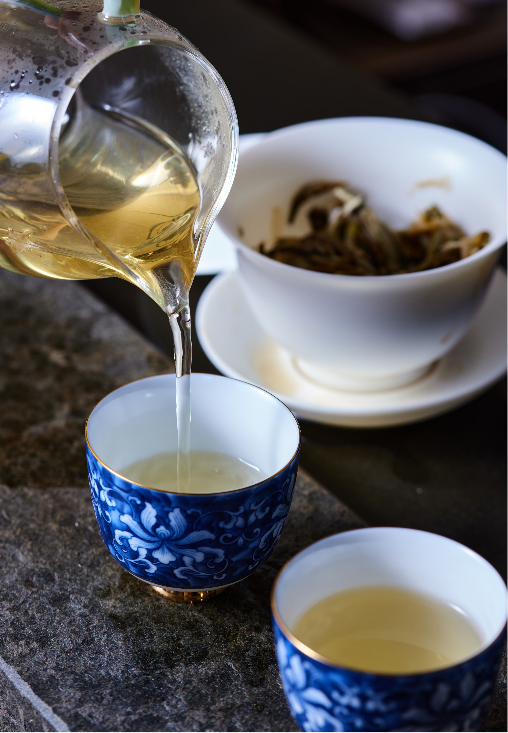 A teapot pouring hot water into a blue and white patterned teacup, with a white bowl containing tea and a bowl of dried tea leaves in the background.