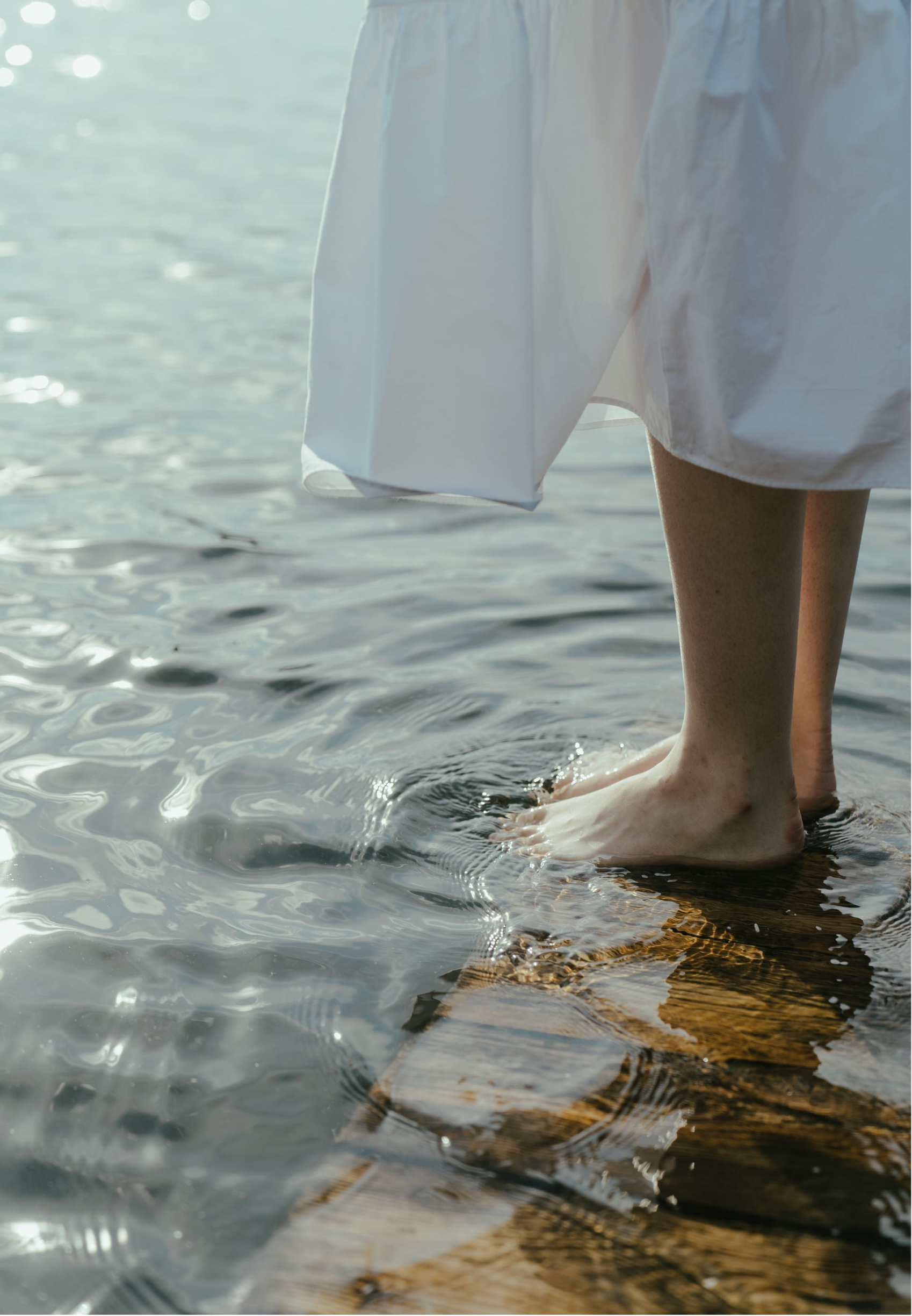 Person standing in shallow water on a wooden dock, wearing a white dress or skirt, with feet partly submerged in water.