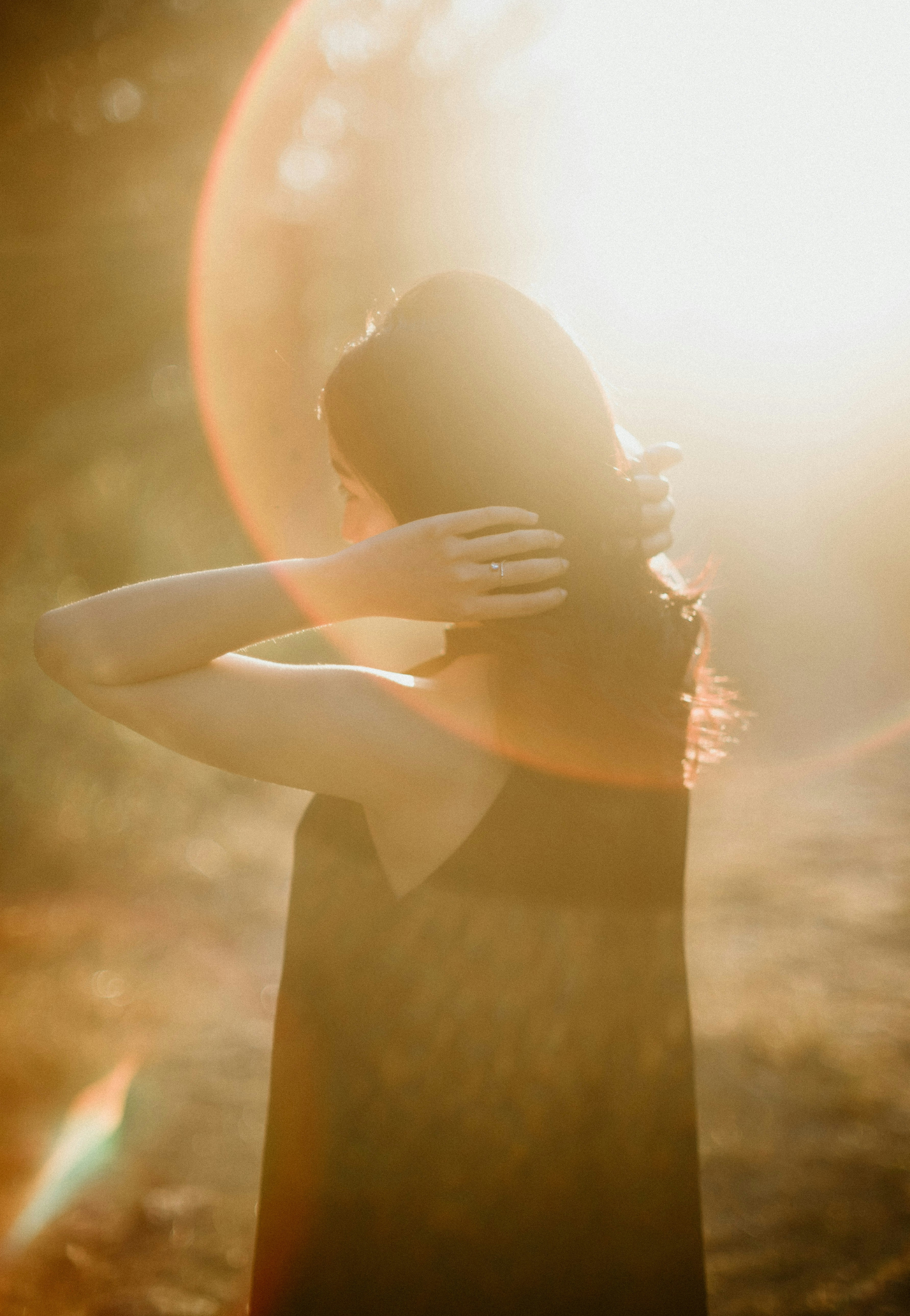 A woman with long hair embracing herself in a natural outdoor setting with sunlight creating a glowing effect.