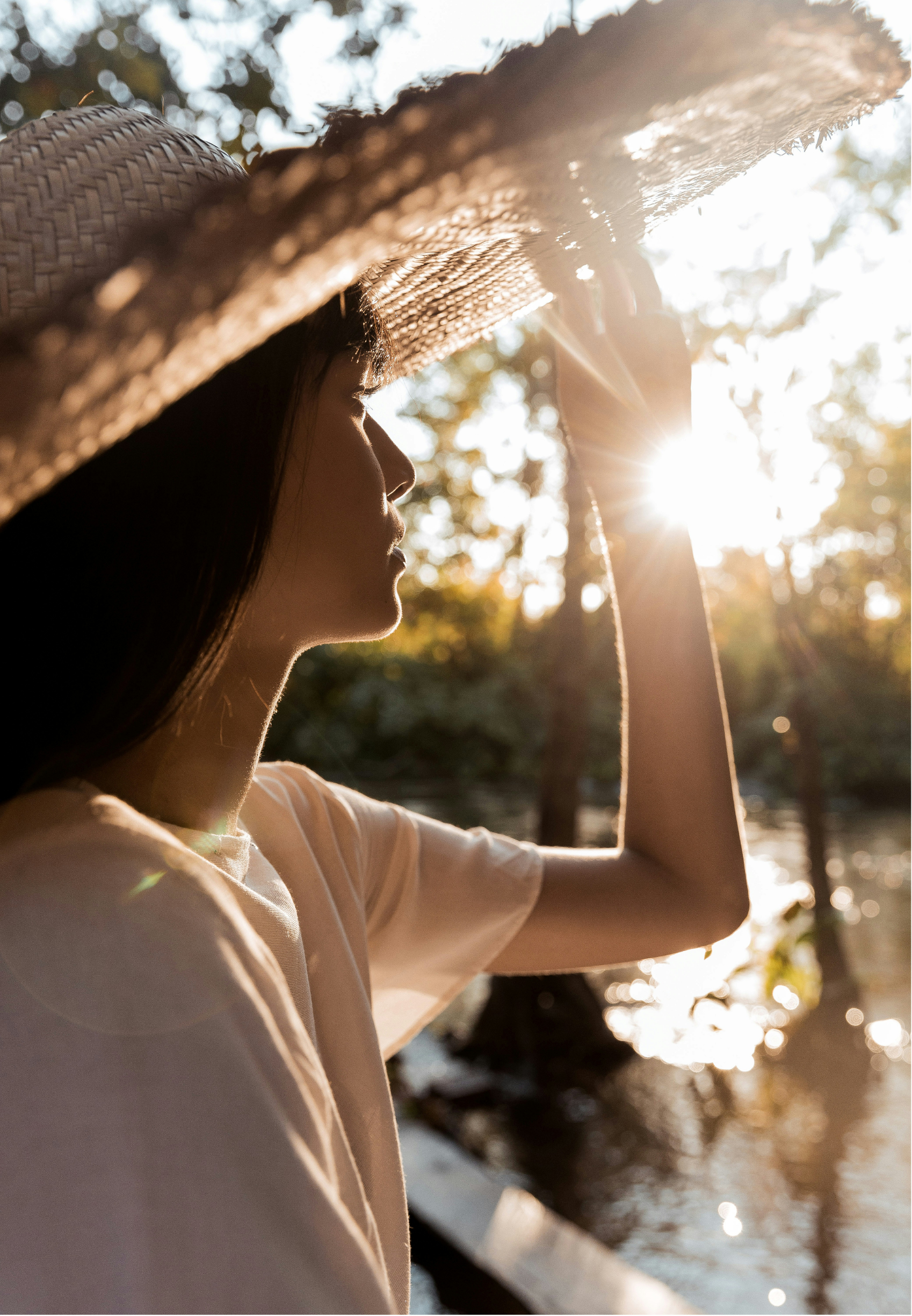 A woman wearing a straw hat and a white shirt standing outdoors at sunset, near water, with sunlight filtering through trees.