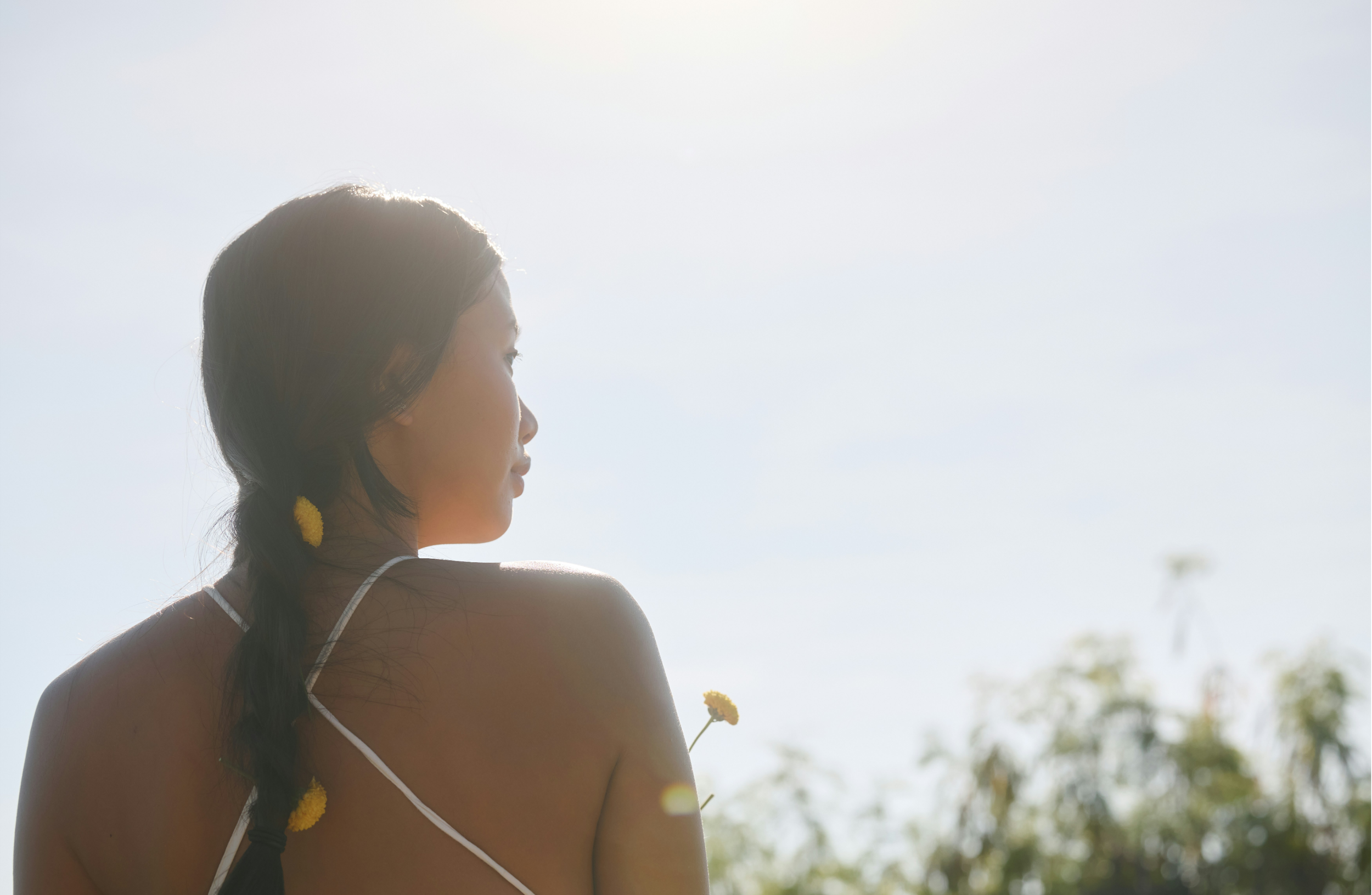 A woman with dark hair tied in a braid decorated with yellow pompoms, gazing thoughtfully to her left outdoors in a sunny setting.