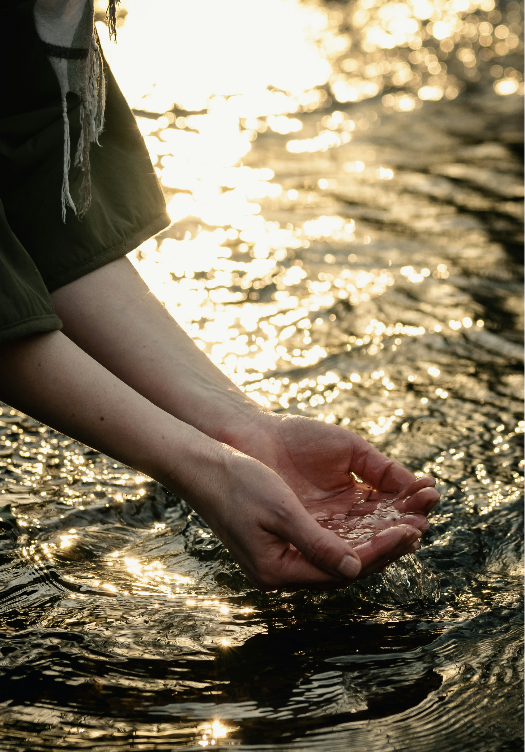 Person's arm and hand submerged in water, cupping water, with sunlight reflecting off the water's surface in the background.
