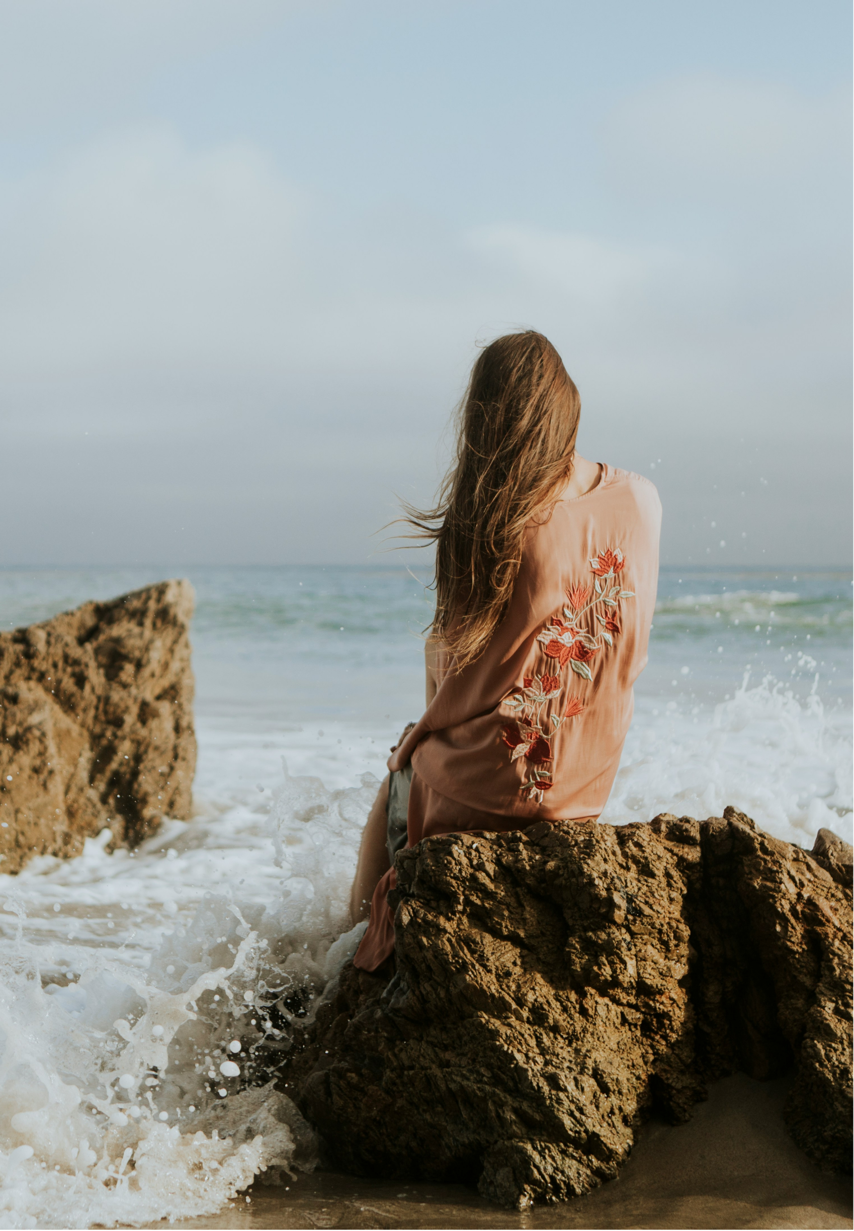 A woman with long hair sitting on a rock by the ocean, facing away, wearing a pink embroidered blouse, waves crashing around rocks.