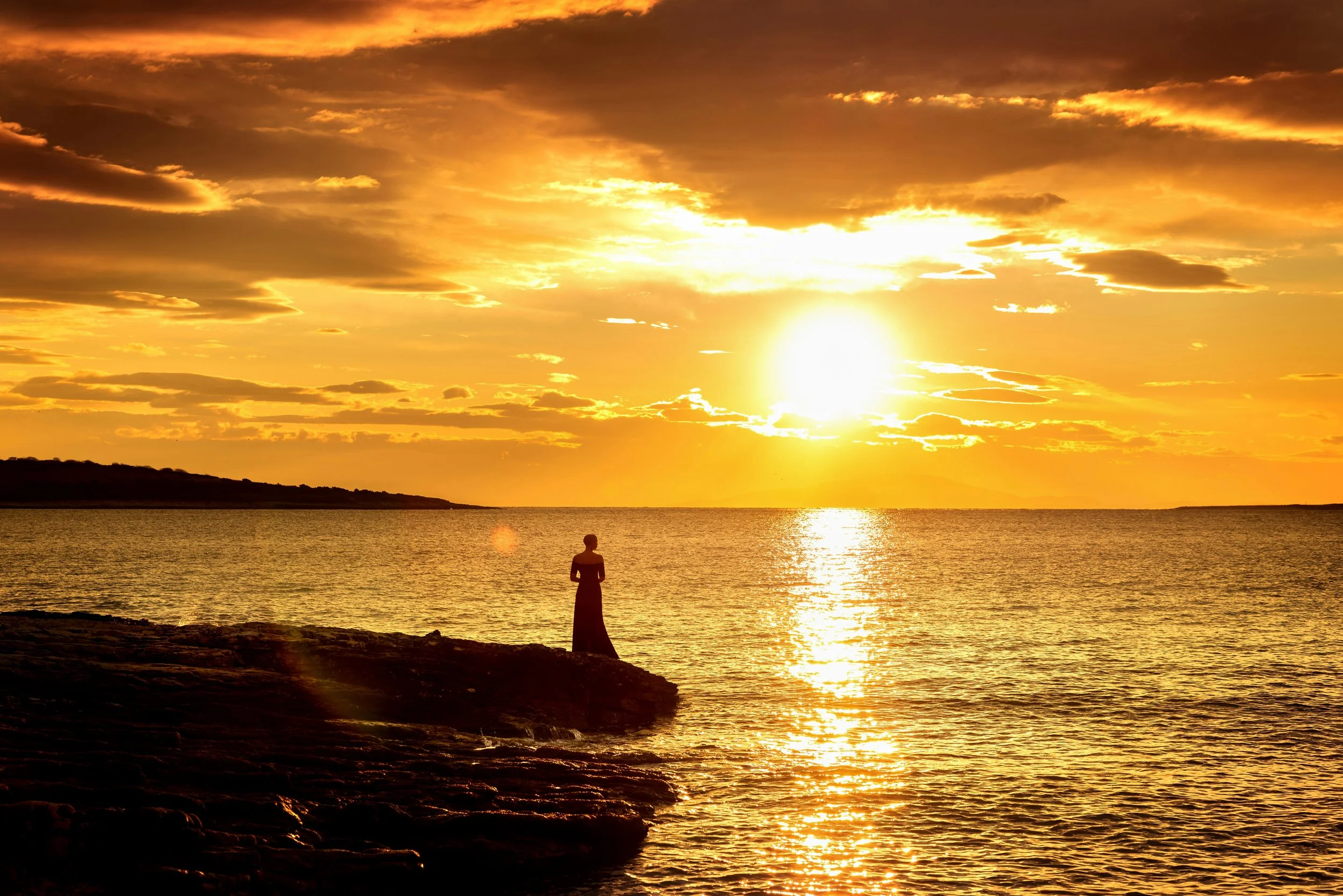 A person standing on a rocky shoreline, silhouetted against a vibrant sunset over a body of water, with the sun partially obscured by clouds.