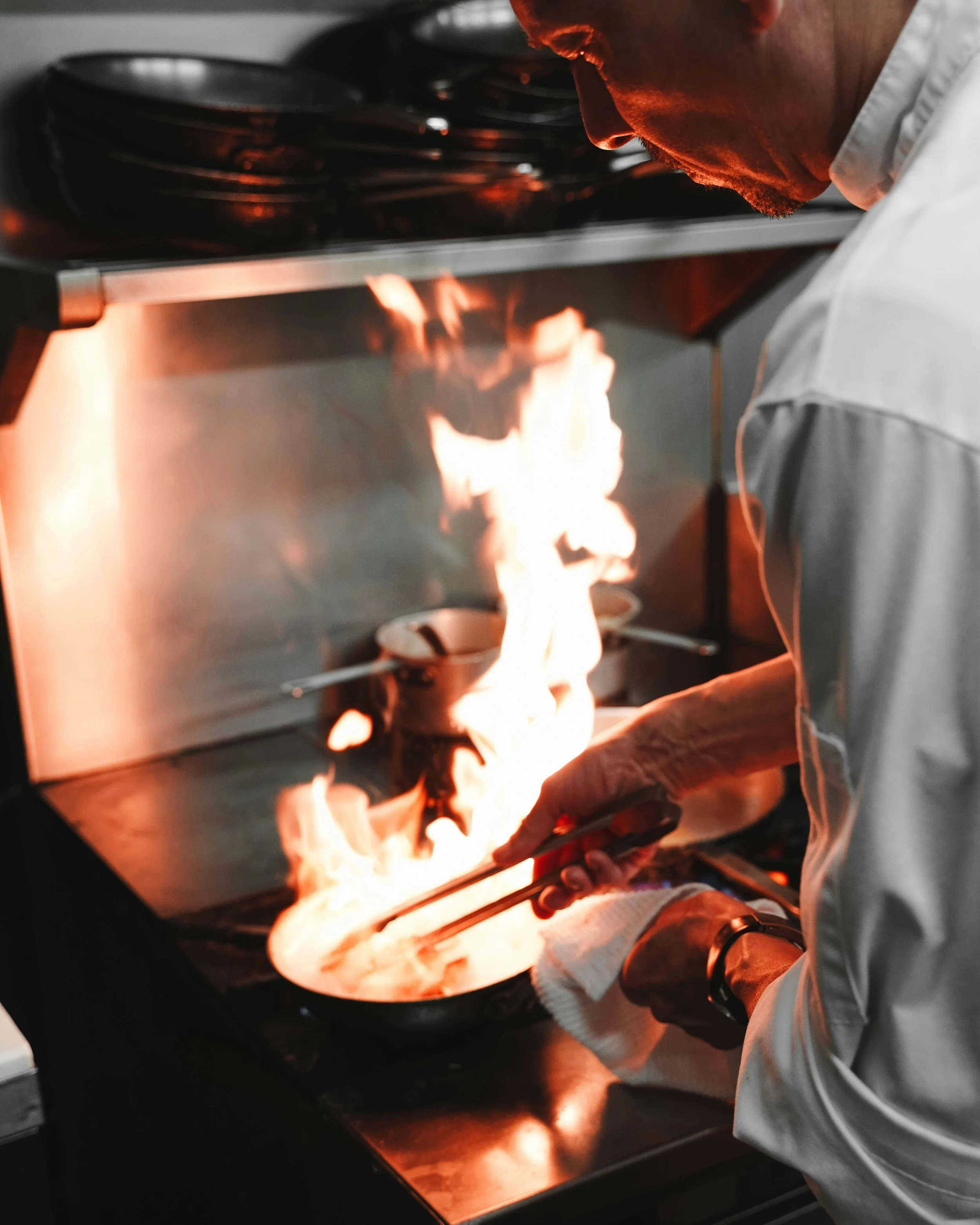 A chef cooking on a stove with flames, wearing a white chef coat, handling a pan with a towel.