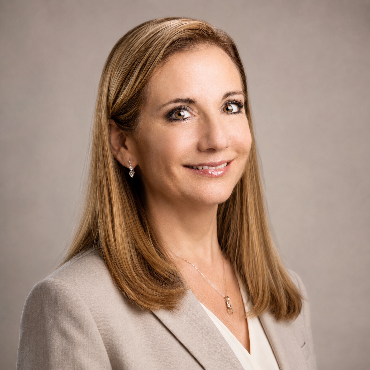 Professional woman with long, straight, light brown hair smiling at the camera, wearing a beige blazer, earrings, and a necklace, against a neutral background.