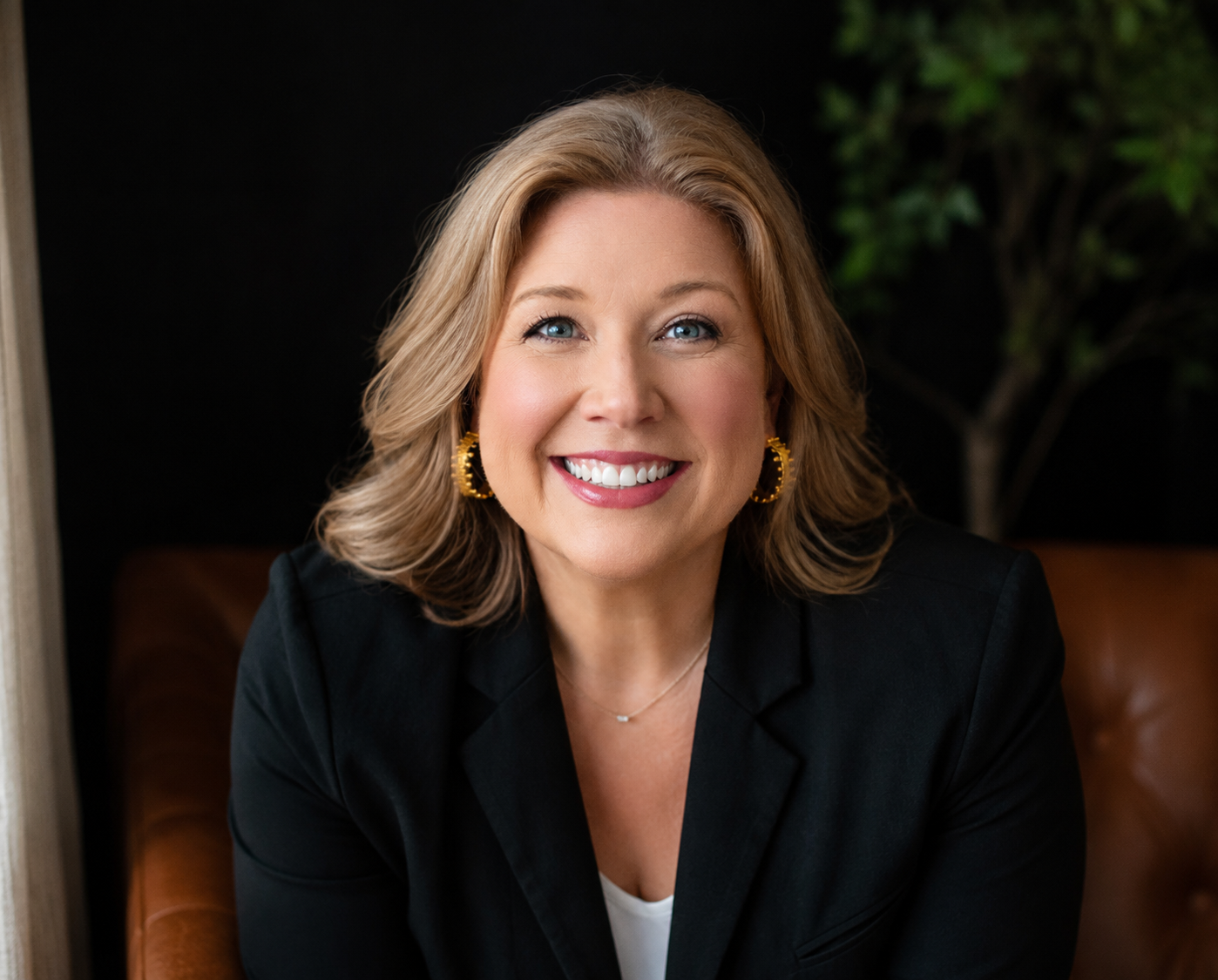 A woman with shoulder-length blonde hair smiling at the camera, wearing a black blazer, gold earrings, and a delicate necklace, sitting on a brown leather chair with a dark background and some greenery.