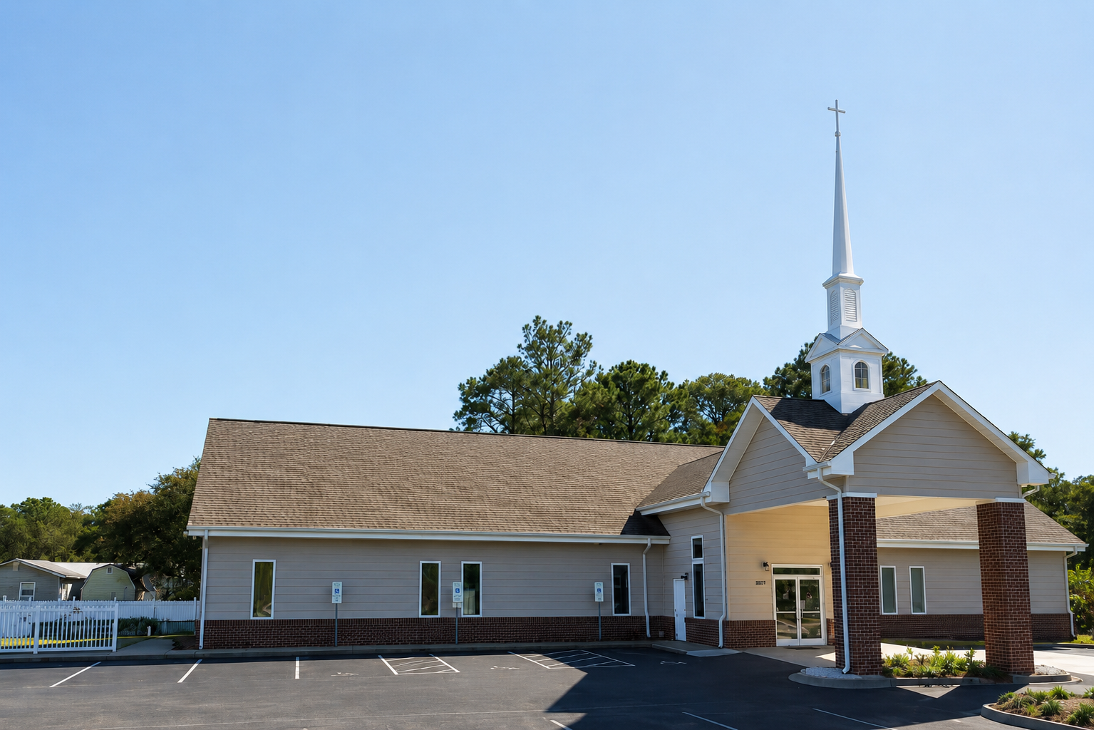 A church building with beige siding, brick accents, and a tall white steeple with a cross, surrounded by an empty parking lot and green trees under a clear blue sky.