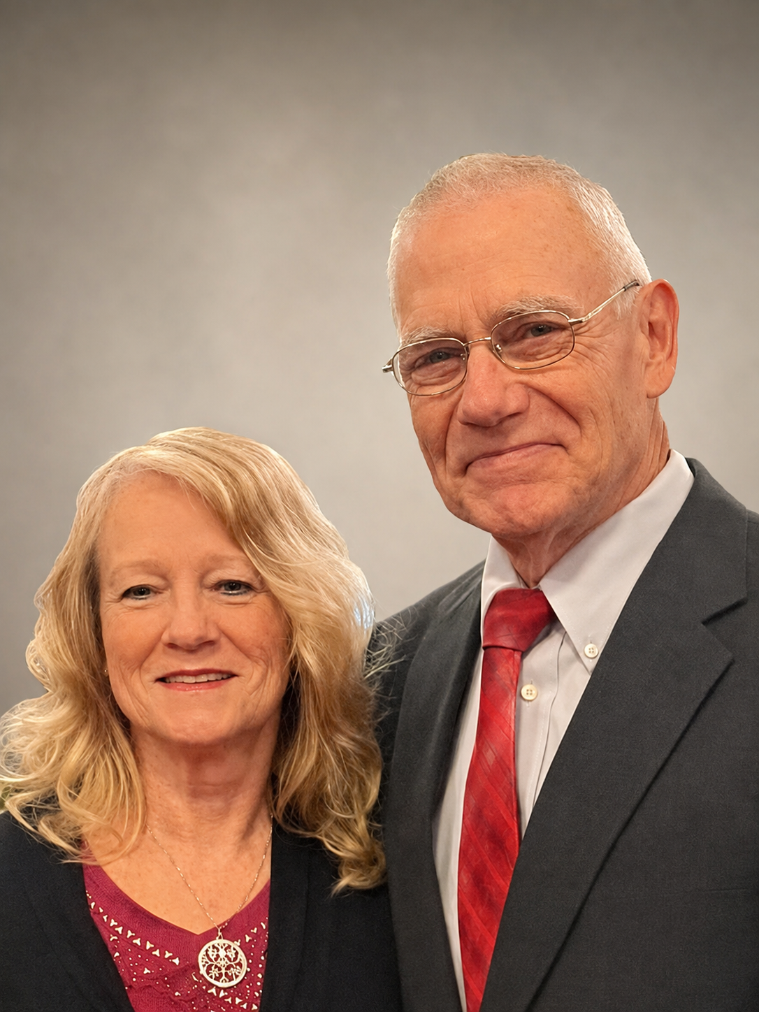 A smiling woman with wavy blond hair wearing a black blazer and a red shirt with a pendant. A smiling man with short gray hair, glasses, wearing a dark suit, white shirt, and red tie, standing beside her against a gray background.