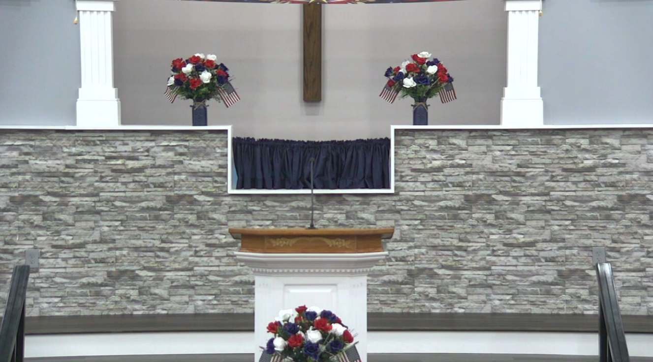 Inside a church, a podium with a microphone, two large flower arrangements with red, white, and blue flowers and small American flags, a cross on the back wall, and a black curtain beneath the cross.