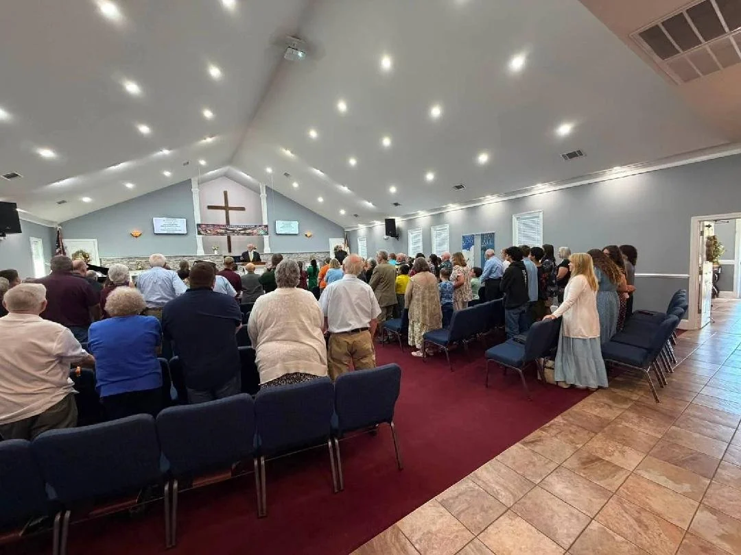 Congregation attending church service in a chapel with a cross on the wall and music screens, some people standing and some sitting.