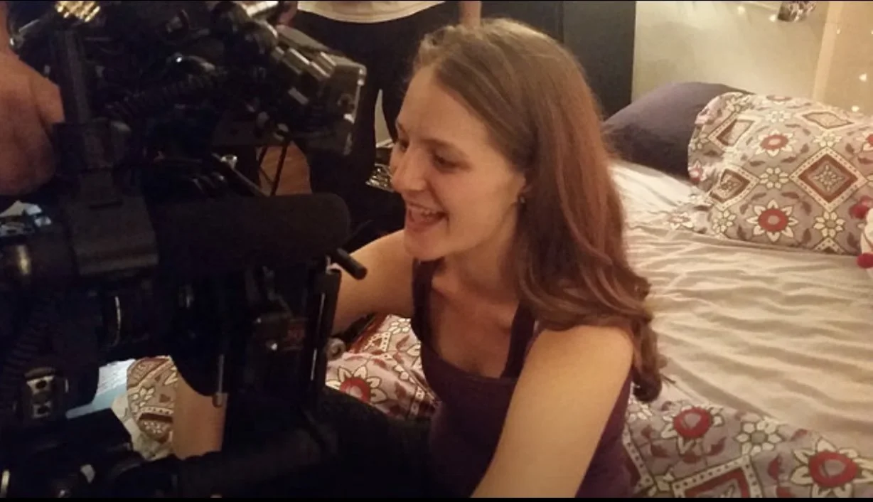A woman with long reddish-brown hair smiling and looking at a film camera on set, sitting on a bed with patterned sheets and pillows.