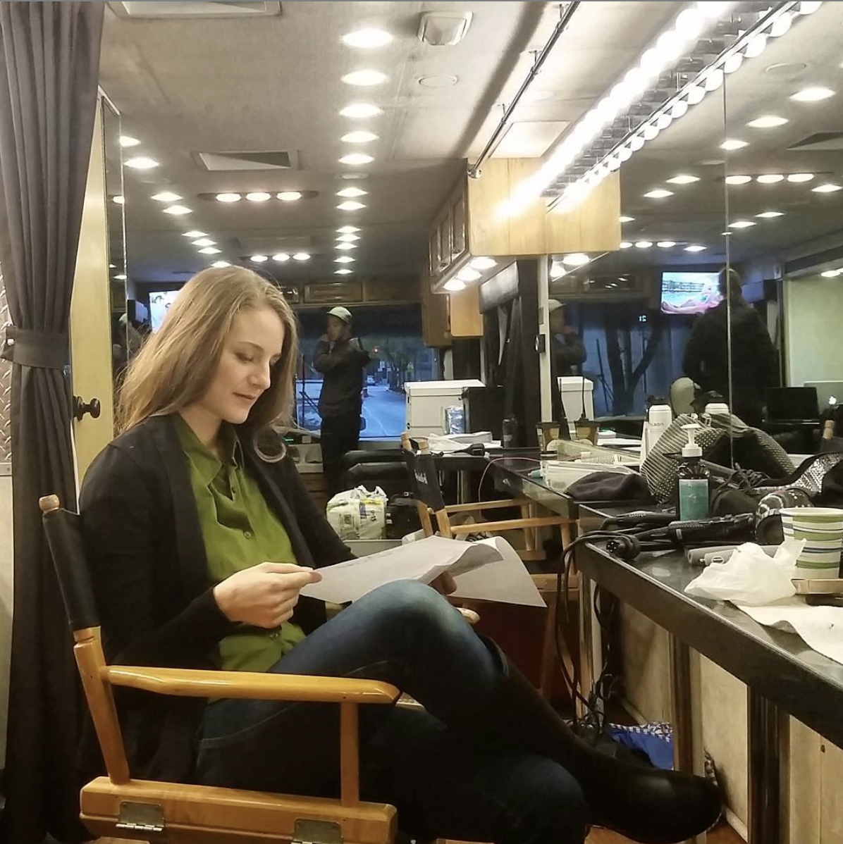 A young woman sitting on a wooden chair, reading a script or papers in a backstage or behind-the-scenes setting with various equipment, bottles, and clutter on a nearby table. A mirror reflects the scene, and another person is visible outside through