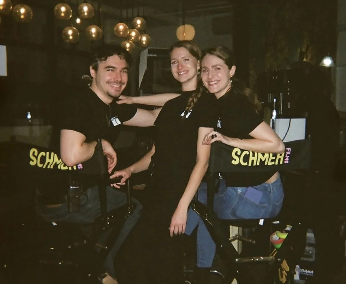 Three young adults, two women and one man, posing together indoors at a bar or club, smiling at the camera, with decorative hanging lights in the background.