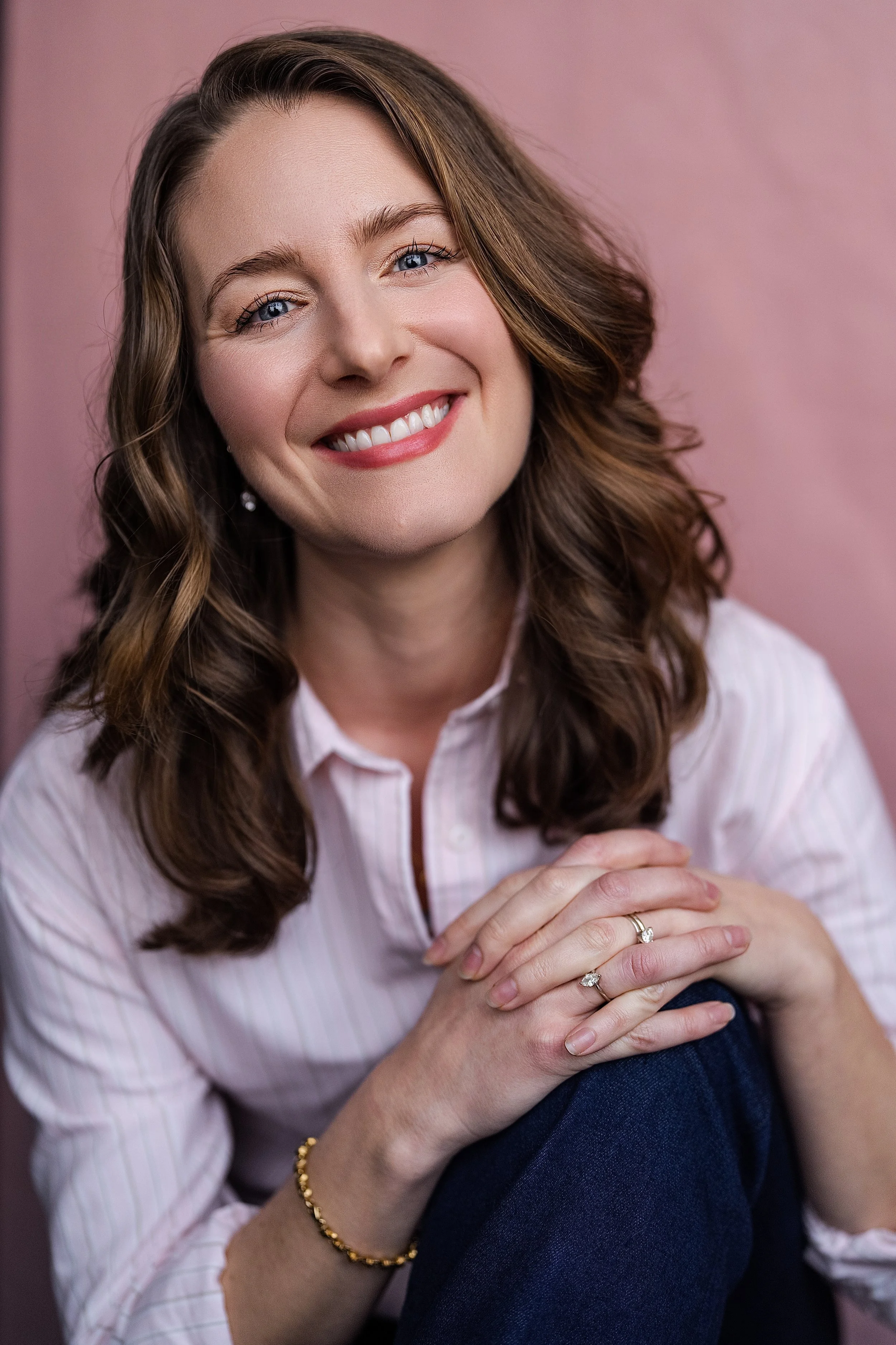 A woman with wavy brown hair, blue eyes, and a bright smile, wearing a white button-up shirt, gold bracelet, and rings, sitting with her hands clasped. The background is pink.
