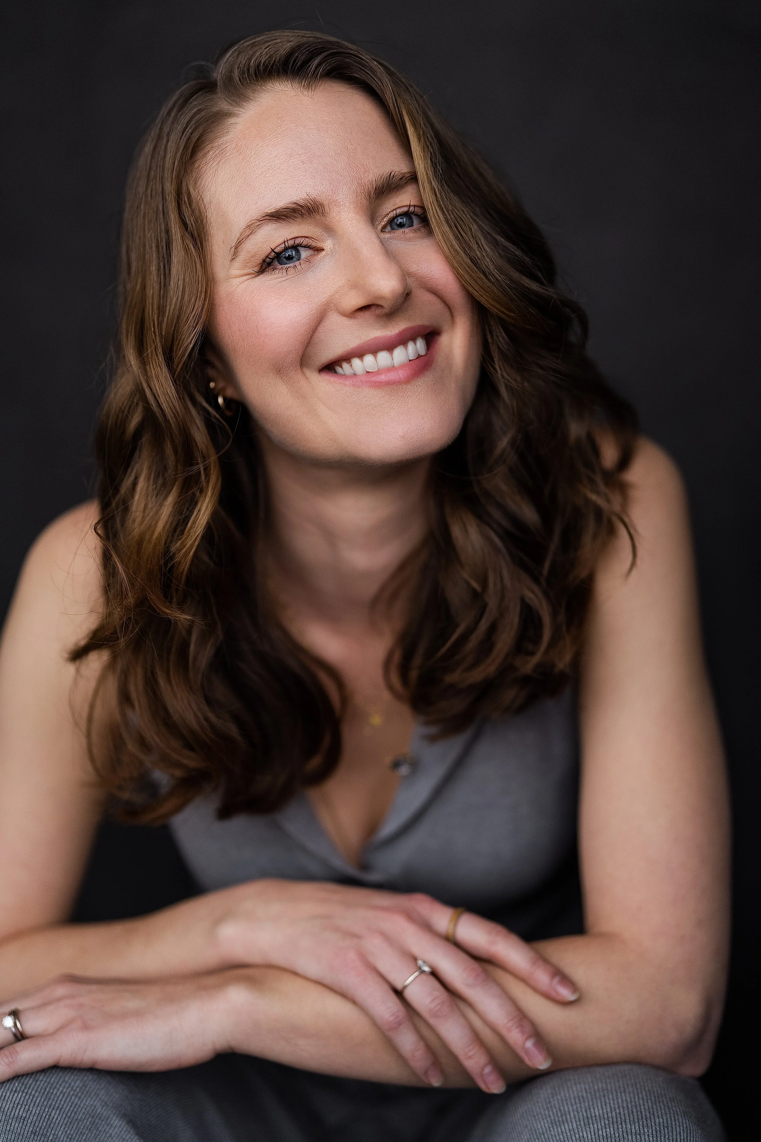 A woman with long wavy brown hair and blue eyes smiling, seated with her arms crossed on her lap against a dark background, wearing a gray top and multiple rings.