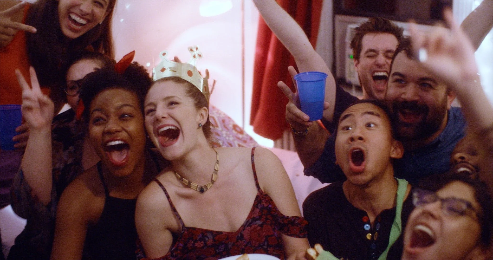 Group of people celebrating, some wearing party hats, with expressions of joy and surprise, in a decorated indoor setting.