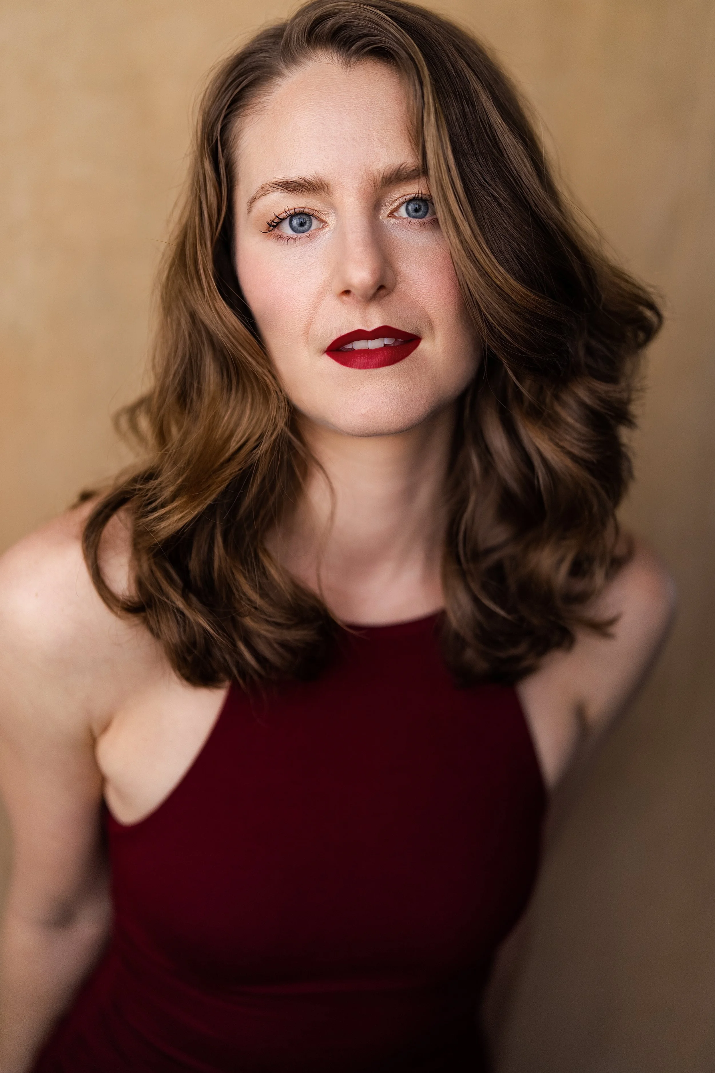 Close-up of a woman with wavy brown hair, blue eyes, and red lipstick wearing a sleeveless burgundy top, posing against a neutral background.