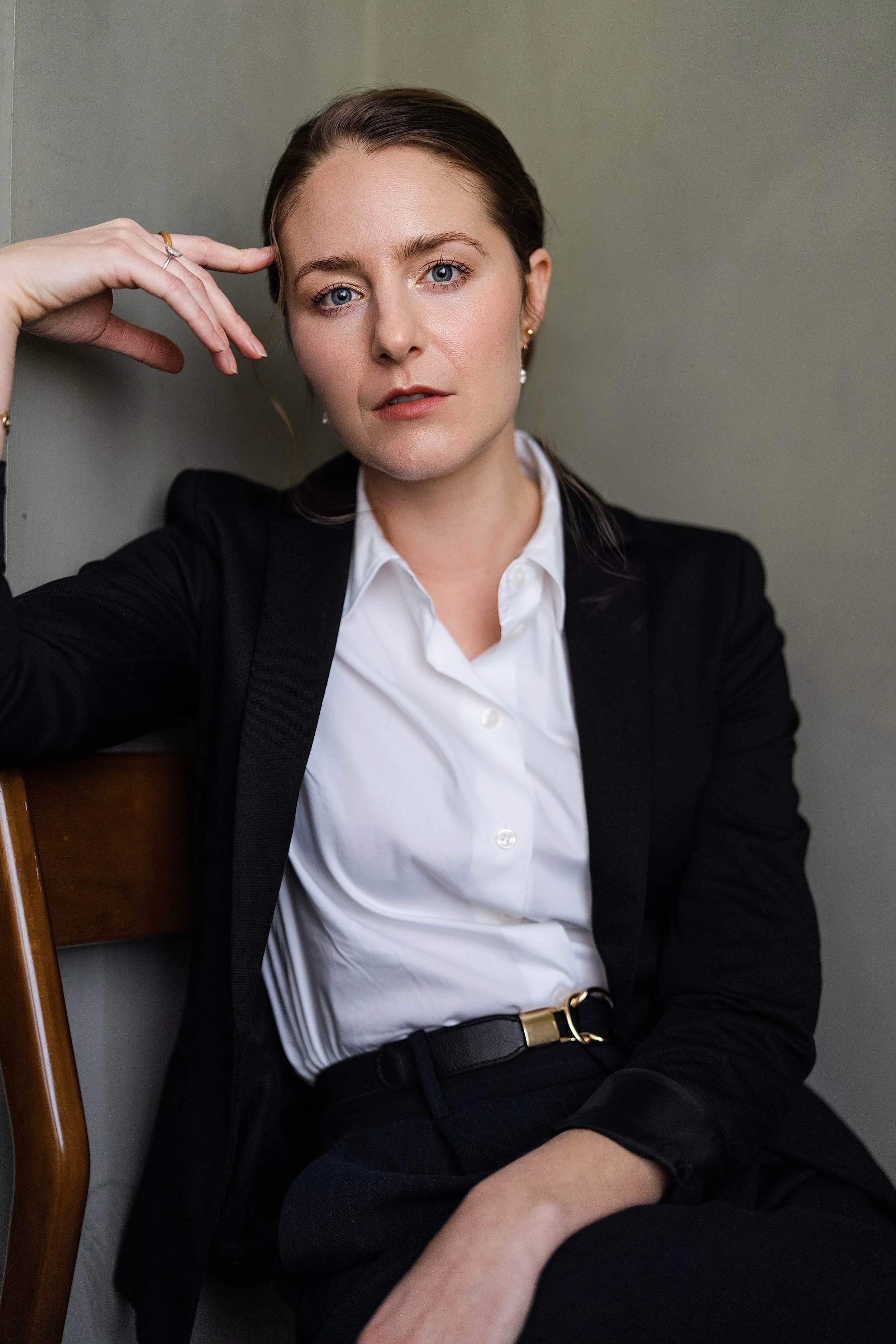 A woman with light skin and blue eyes, wearing a black blazer and white shirt, sitting with her hand resting on her head, and a neutral expression. She has earrings and a ring.