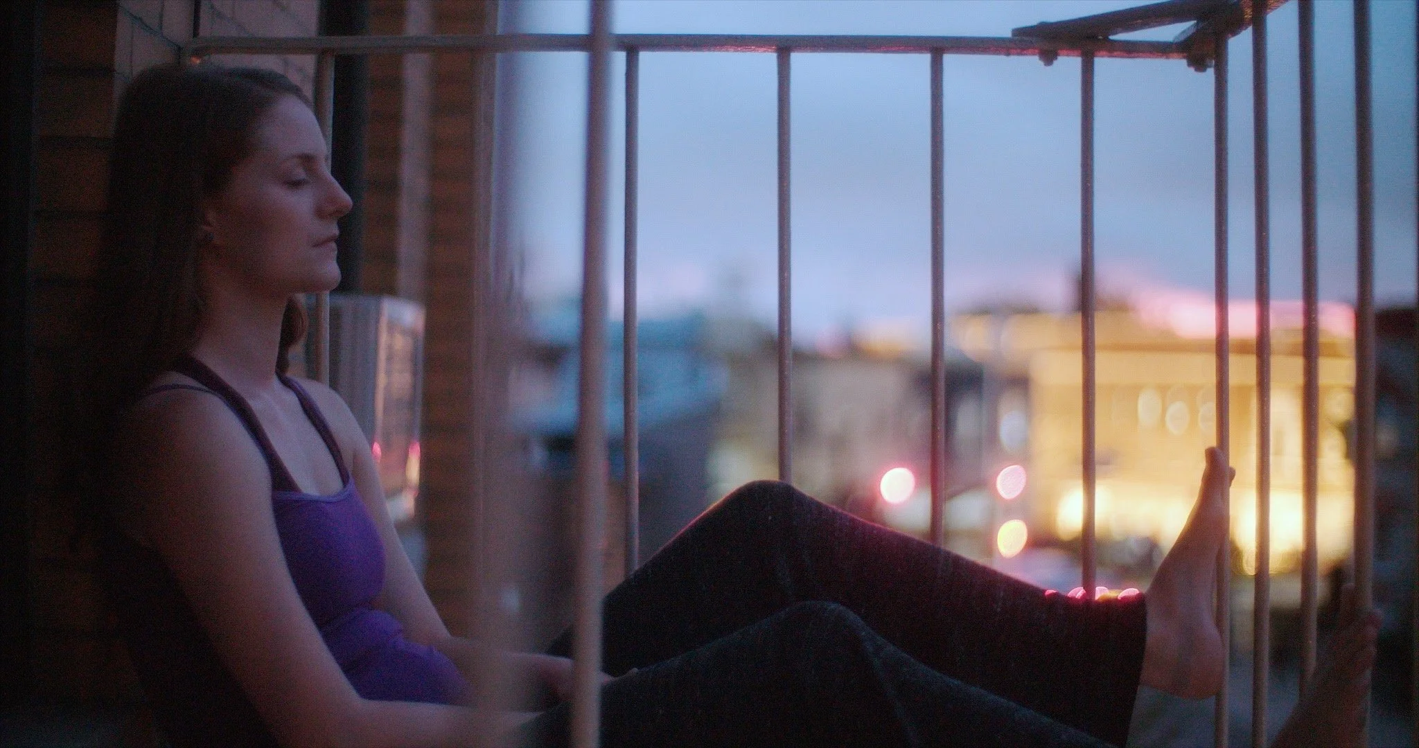 A young woman with long brown hair sitting on a balcony at dusk, holding a cell phone, with city lights and a blue sky in the background.