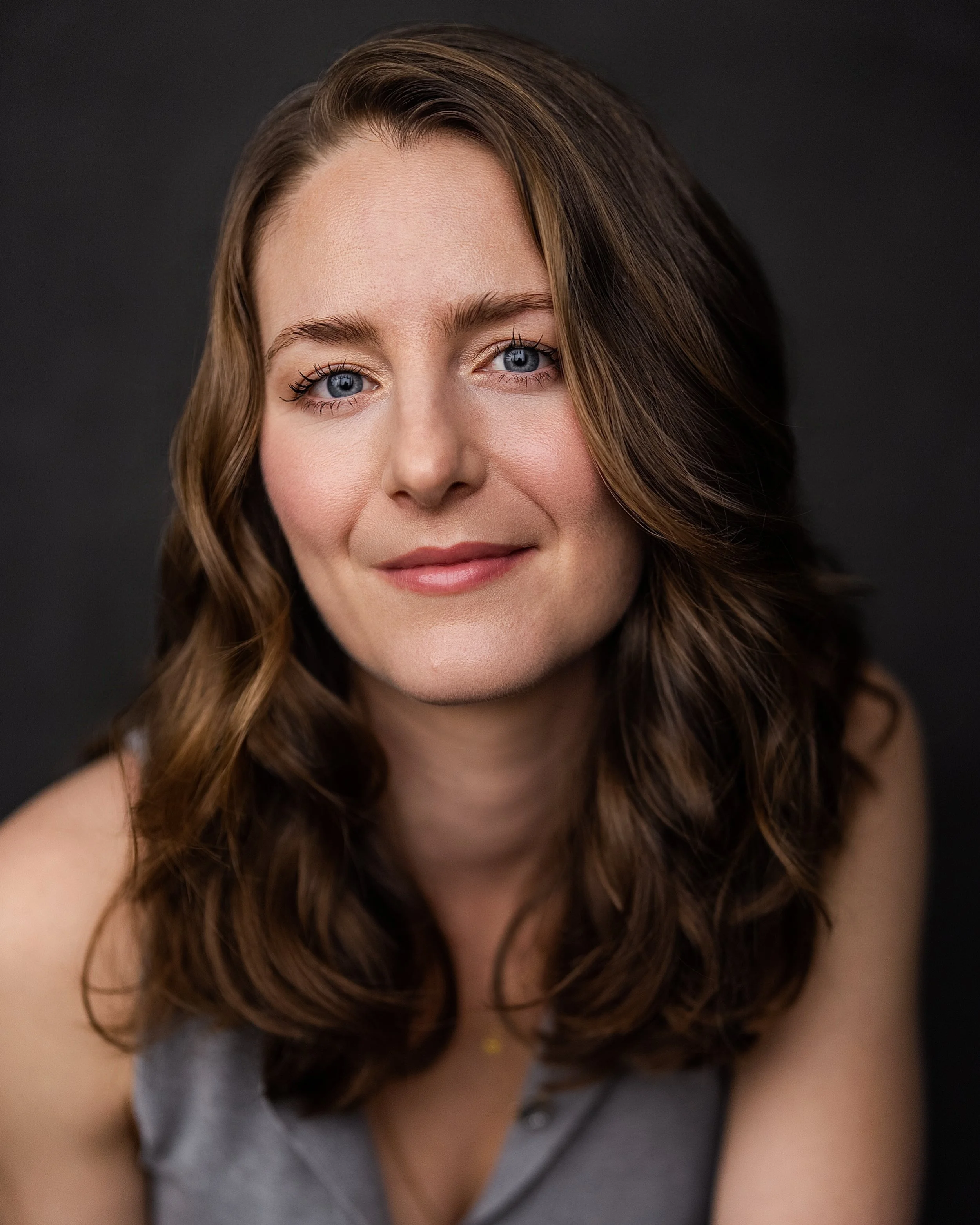 Close-up of a woman with long wavy brown hair, blue eyes, and a gentle smile, wearing a gray top, against a dark background.