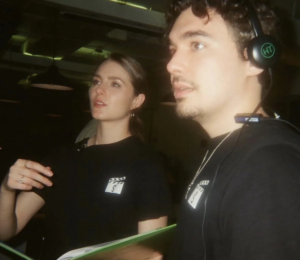 Two young adults, a woman and a man, are in a dimly lit indoor setting, possibly a nightclub or event space. The woman has dark hair styled back and is wearing a black shirt, while the man has curly hair, is wearing headphones, and a black shirt. He 