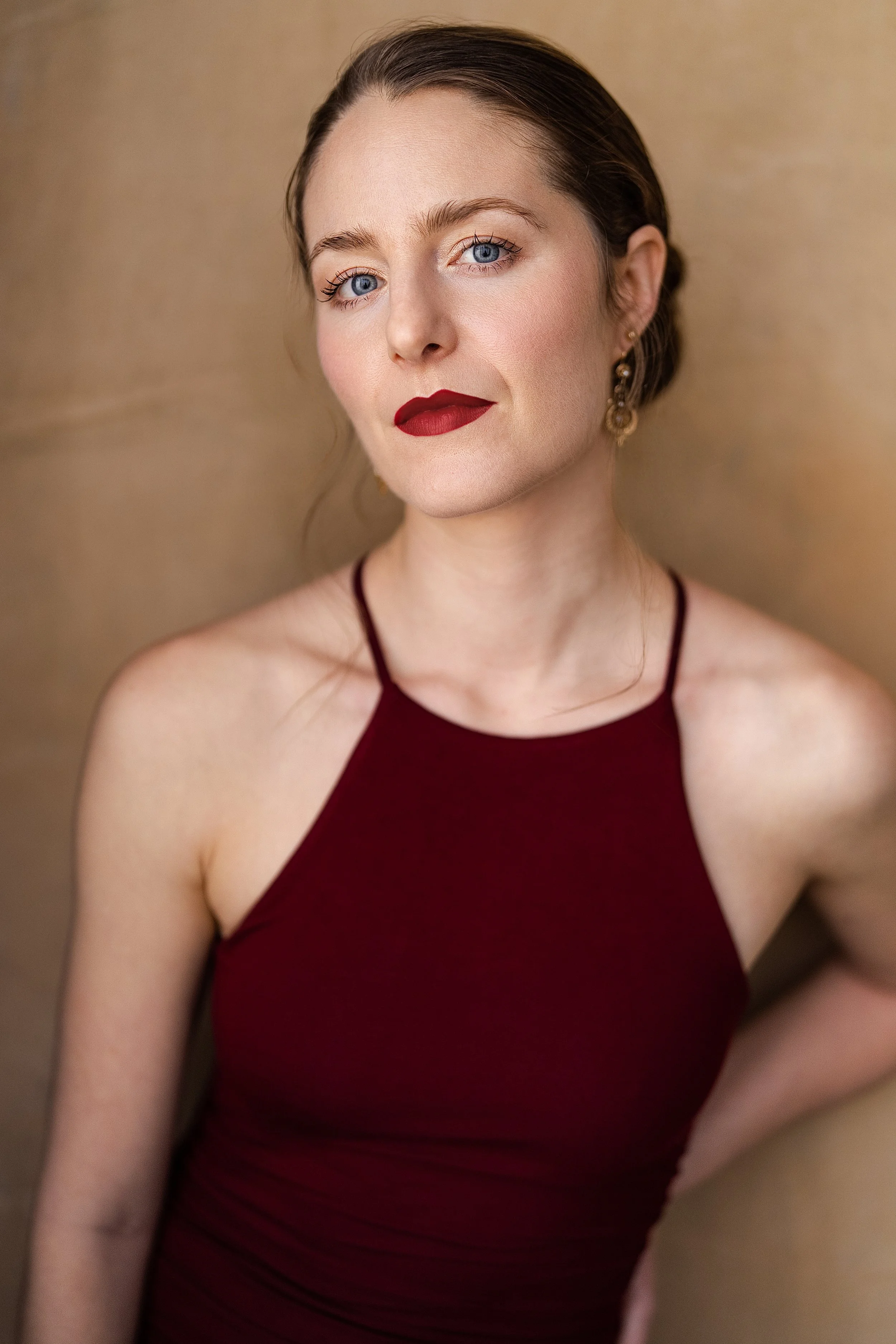 A woman with blue eyes, fair skin, dark brown hair styled in a loose updo, wearing red lipstick, gold earrings, and a dark red halter top against a neutral background.