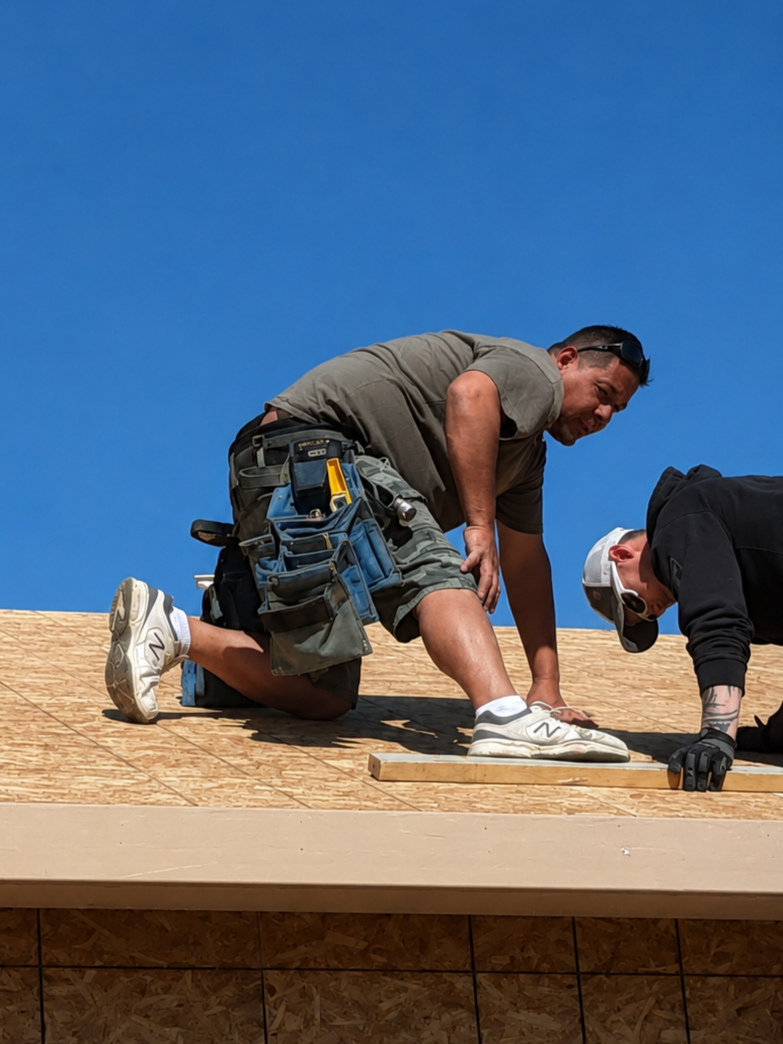 Two construction workers installing roof sheathing on a building under a clear blue sky.