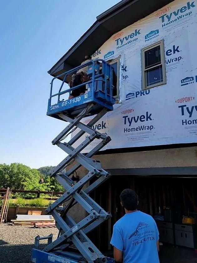 A construction worker standing in a blue scissor lift working on the exterior wall of a house that is covered with Tyvek HomeWrap. Another person in a light blue shirt is standing underneath, watching or assisting. The house has two small windows and