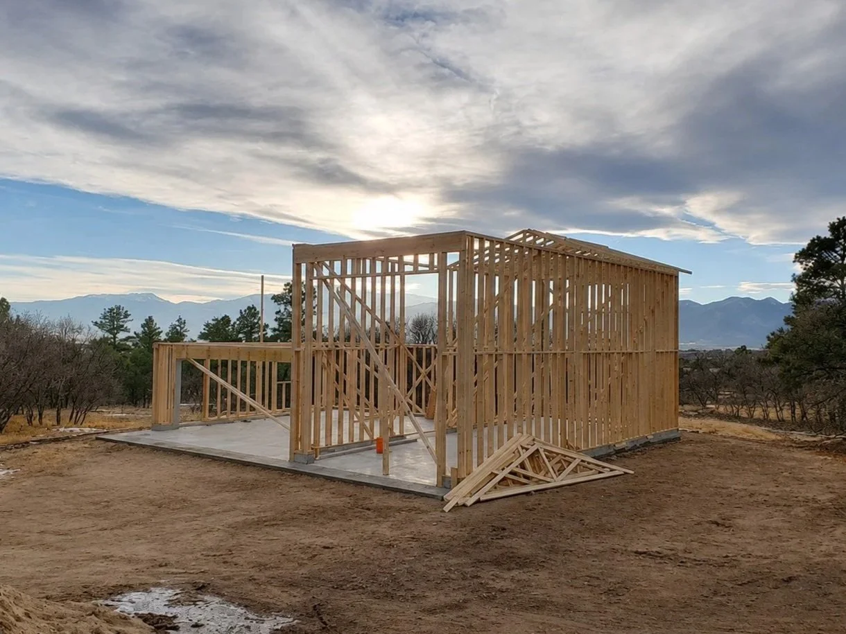 Wooden framing structure of a house under construction on a concrete foundation, with mountains and cloudy sky in the background.