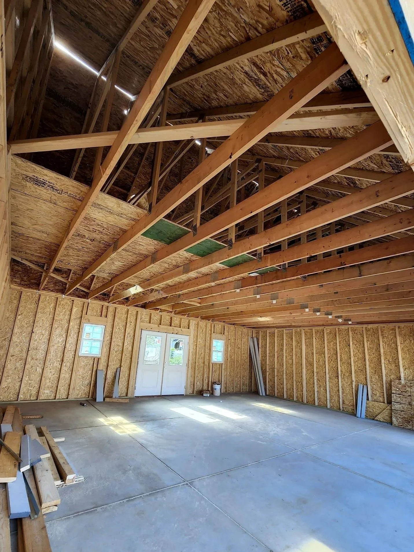 Interior of a building under construction, showing exposed wooden framing, a concrete floor, and part of the roof structure with OSB sheathing. There are two small windows, a door, and construction materials and tools inside.