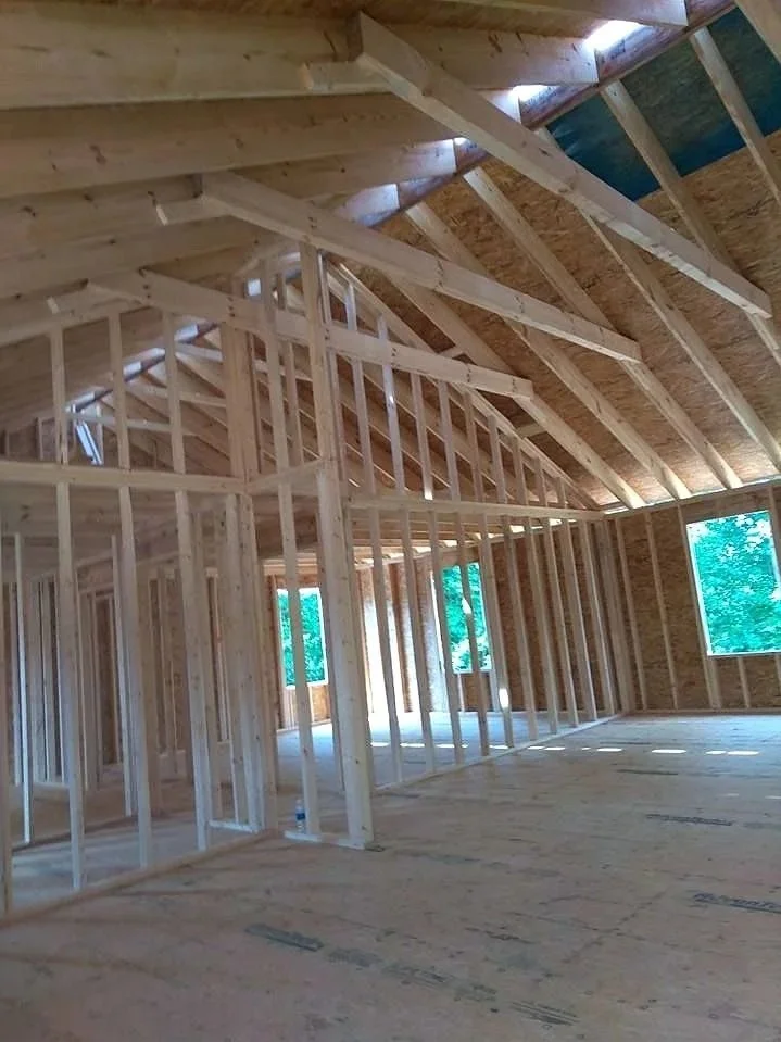 Interior of a house under construction, showing wooden framing and exposed ceiling supports with no drywall or finished surfaces.