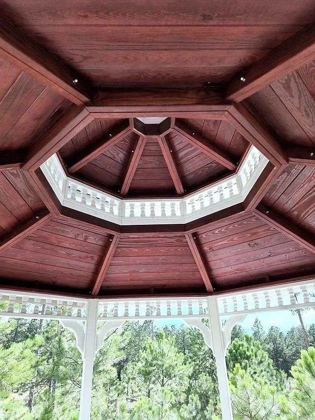 Looking up at a wooden octagonal gazebo ceiling with a white balustrade around the opening and green trees visible outside.
