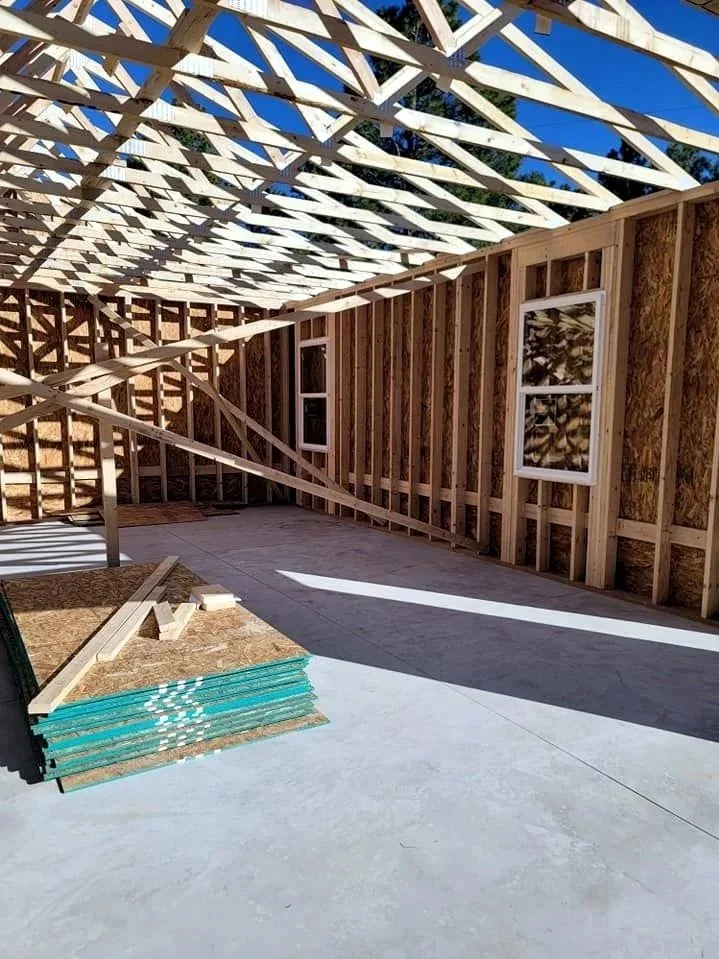 Interior view of a building under construction with exposed wooden framing, a partially assembled roof truss system, and two windows on the wall. There are construction materials on the floor and sunlight casting shadows inside.