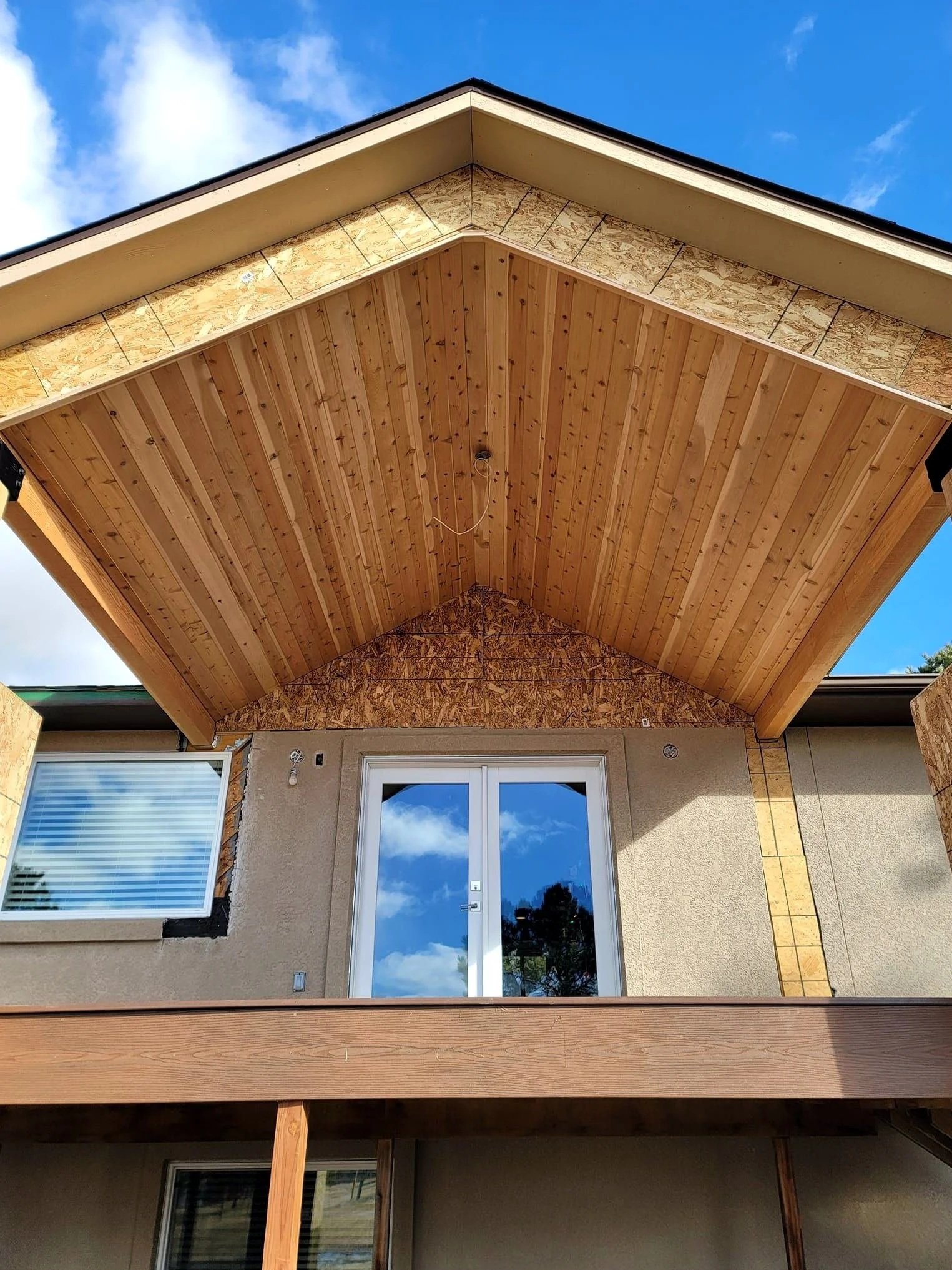 Exterior view of a house with a new wooden porch ceiling and unfinished construction area beneath, showing support beams and construction materials, against a background of a blue sky with clouds.